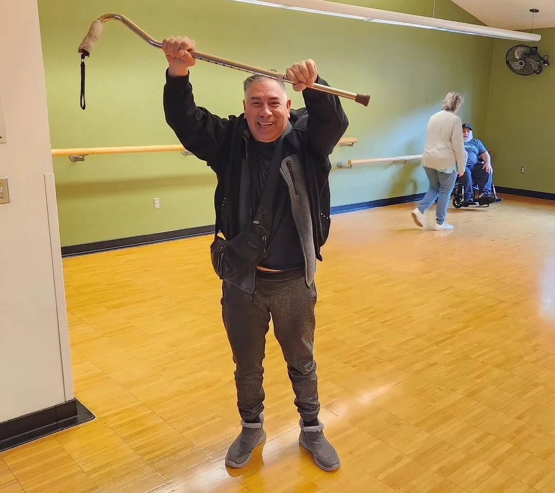 A man smiling and holding a crutch over his head inside a room with a wooden floor, lime green walls, and a mirror.