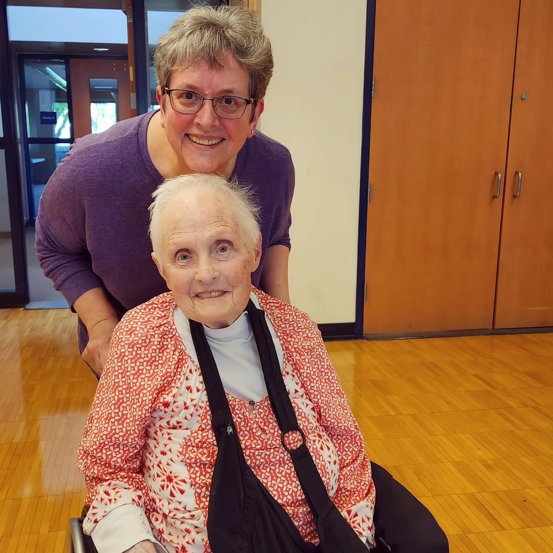 A woman with short gray hair and glasses standing behind an elderly woman sitting in a wheelchair, both smiling at the camera in a room with wooden floors and doors.
