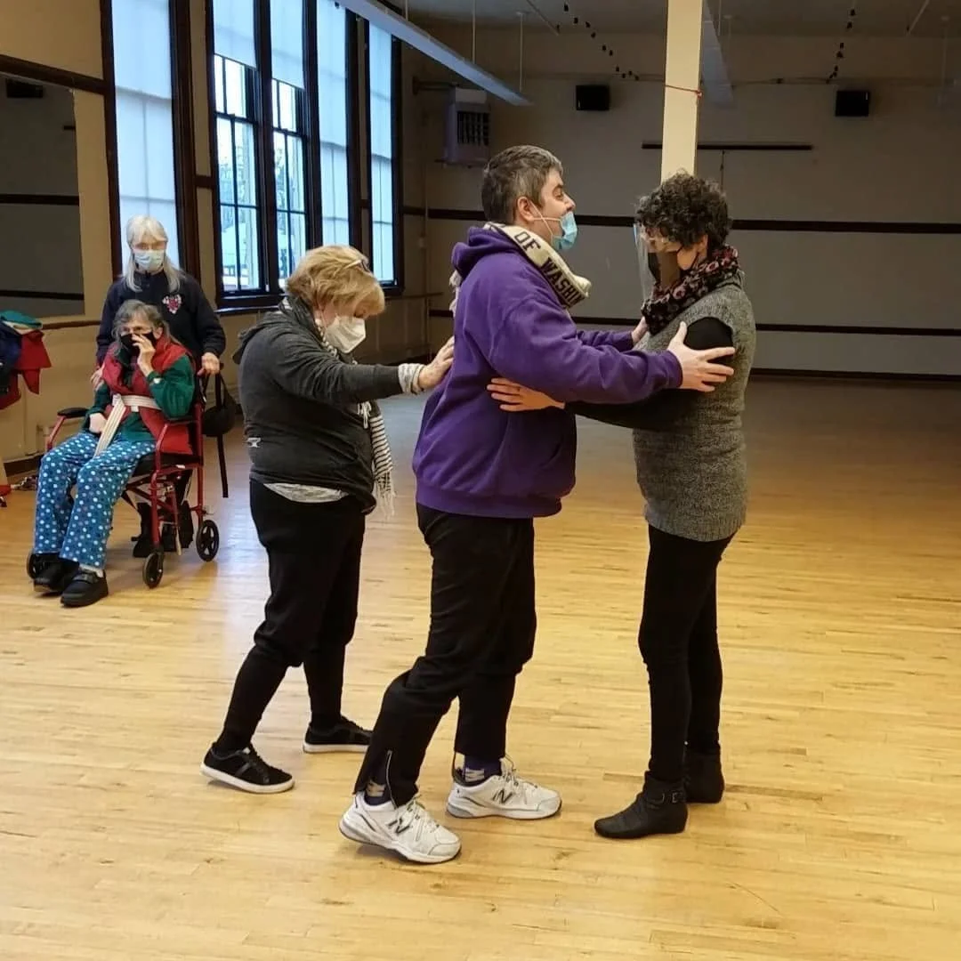 Four people, three women and one man, are dancing in a large room with wooden floors and big windows. The man and one woman are facing each other, holding each other's arms, while the other two women stand nearby, with one touching the shoulder of the woman facing the man. All are wearing face masks.