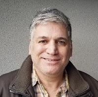Portrait of a smiling middle-aged man with gray hair, wearing a dark coat over a plaid shirt against a plain background.