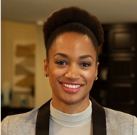 A woman smiling with her hair in an updo, wearing a gray blazer and white top, in an indoor setting.