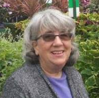 A smiling woman with gray hair and glasses, wearing a gray sweater over a purple top, standing outdoors with greenery in the background.
