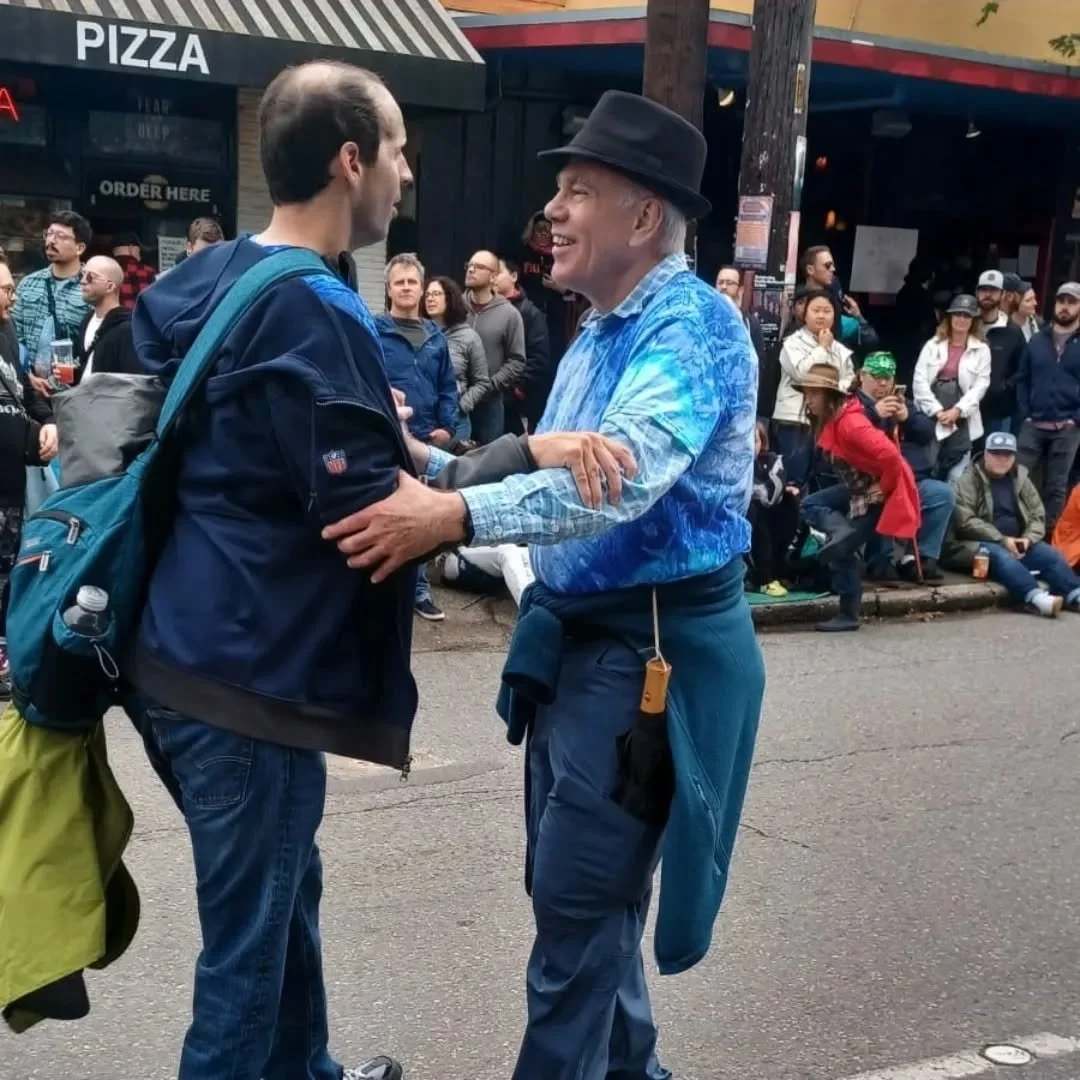 Two men smiling and holding hands on a busy city street, with people in the background, a pizza place sign, and food vendors.