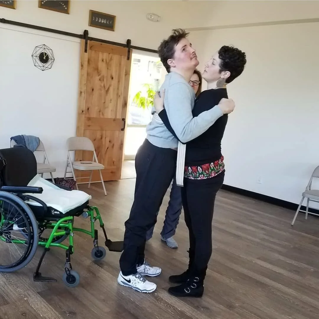 Two women embrace in a room with wooden floors and a partially open door, with a wheelchair nearby and several chairs against the wall.