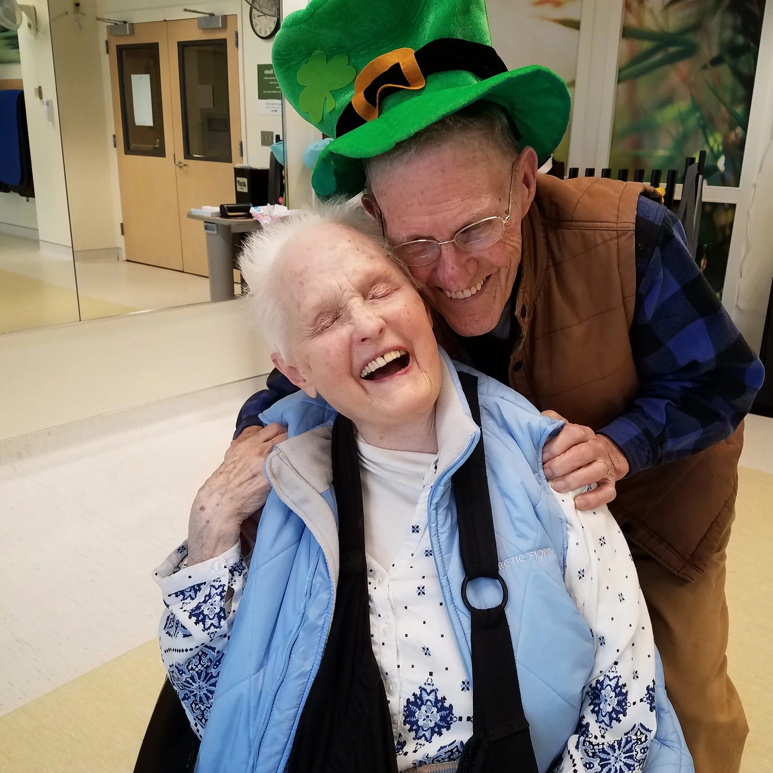 An elderly woman wearing a light blue vest, white shirt, and glasses, hugging a man wearing a green hat decorated with a four-leaf clover and a black bow, in an indoor setting with yellow and cream walls and a mirrored closet.