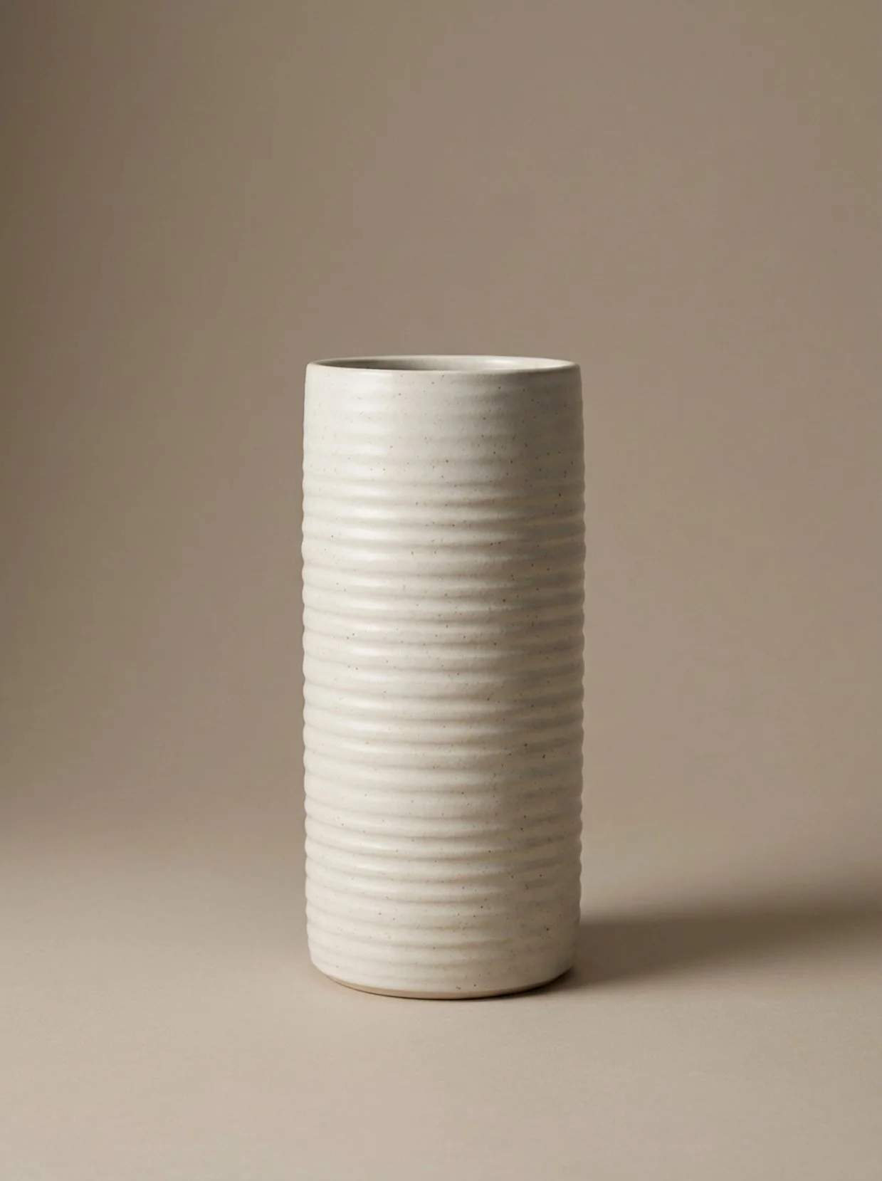 White, ribbed ceramic vase with a cylindrical shape, placed on a plain beige surface with a beige background.