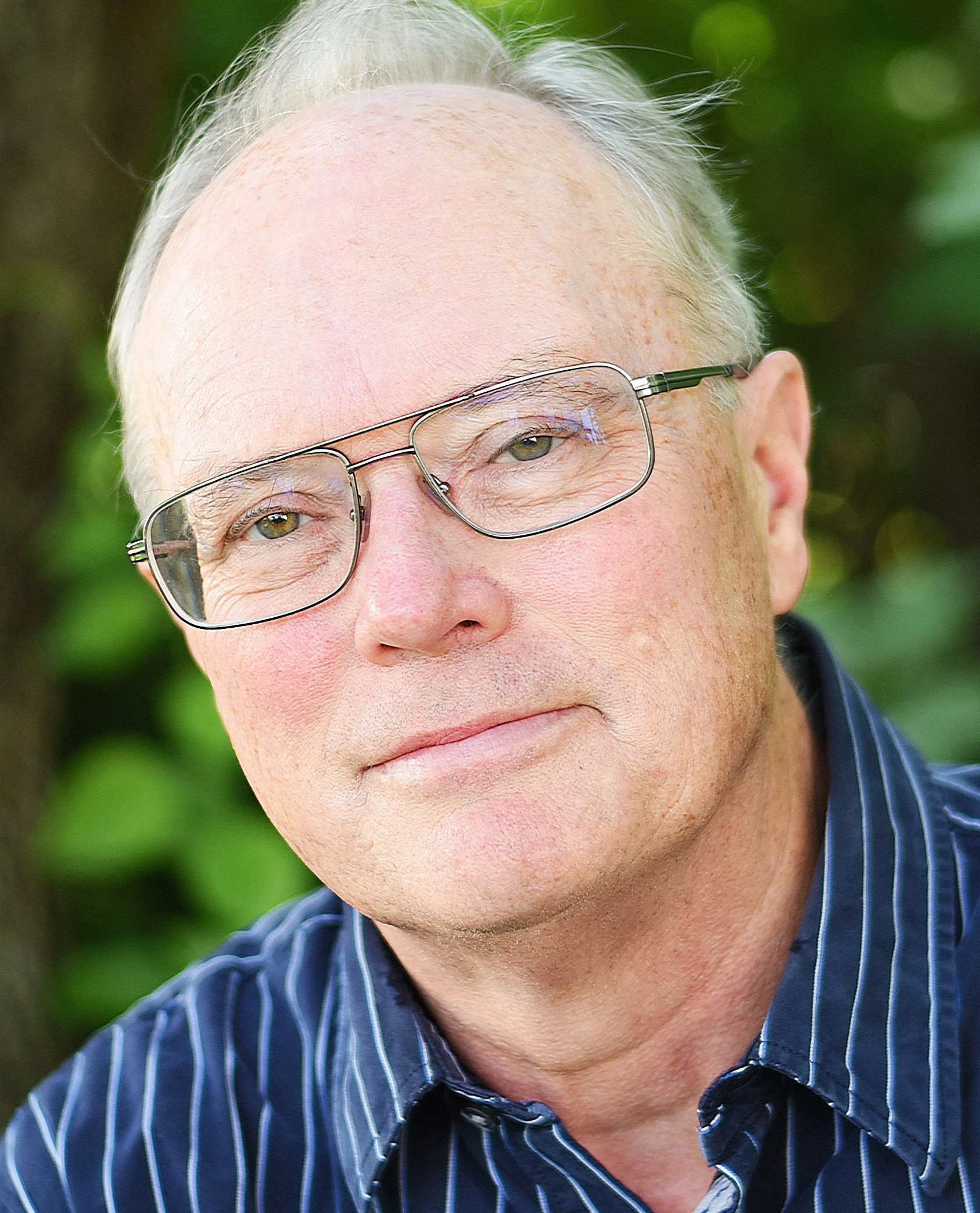 Douglas Rooks has light skin, wears glasses, and has gray hair, dressed in a dark blue striped shirt. He is outdoors with foliage in the background.