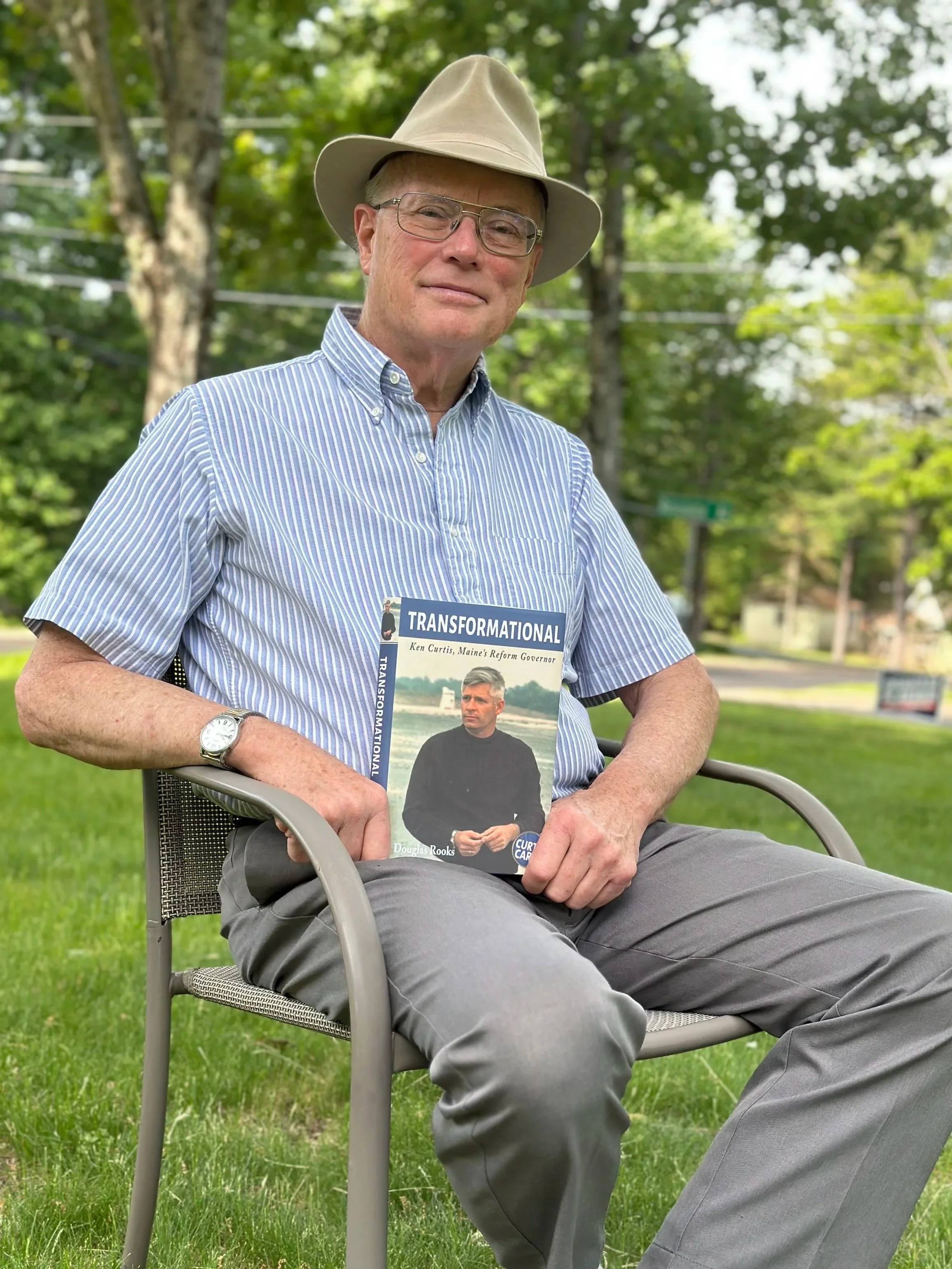 Douglas Rooks sits outdoors on a green lawn, holding a book titled 'Transformational,' wearing a blue and white striped shirt, gray pants, a beigte hat, and glasses.