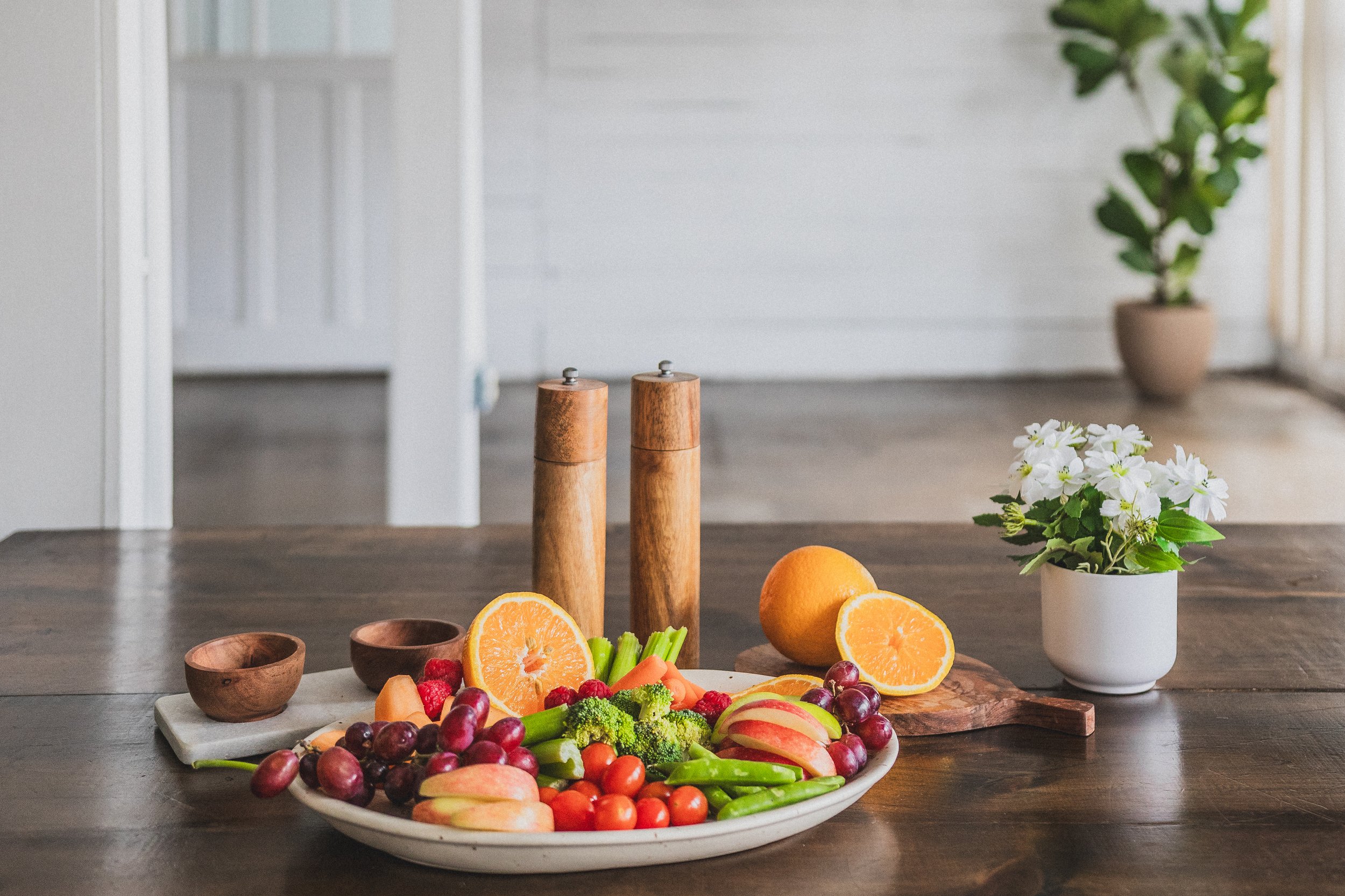 A fruit and vegetable platter with oranges, grapes, apples, celery, broccoli, cherries, and strawberries on a white plate on a wooden table, with a white flower pot and potted plant in the background.