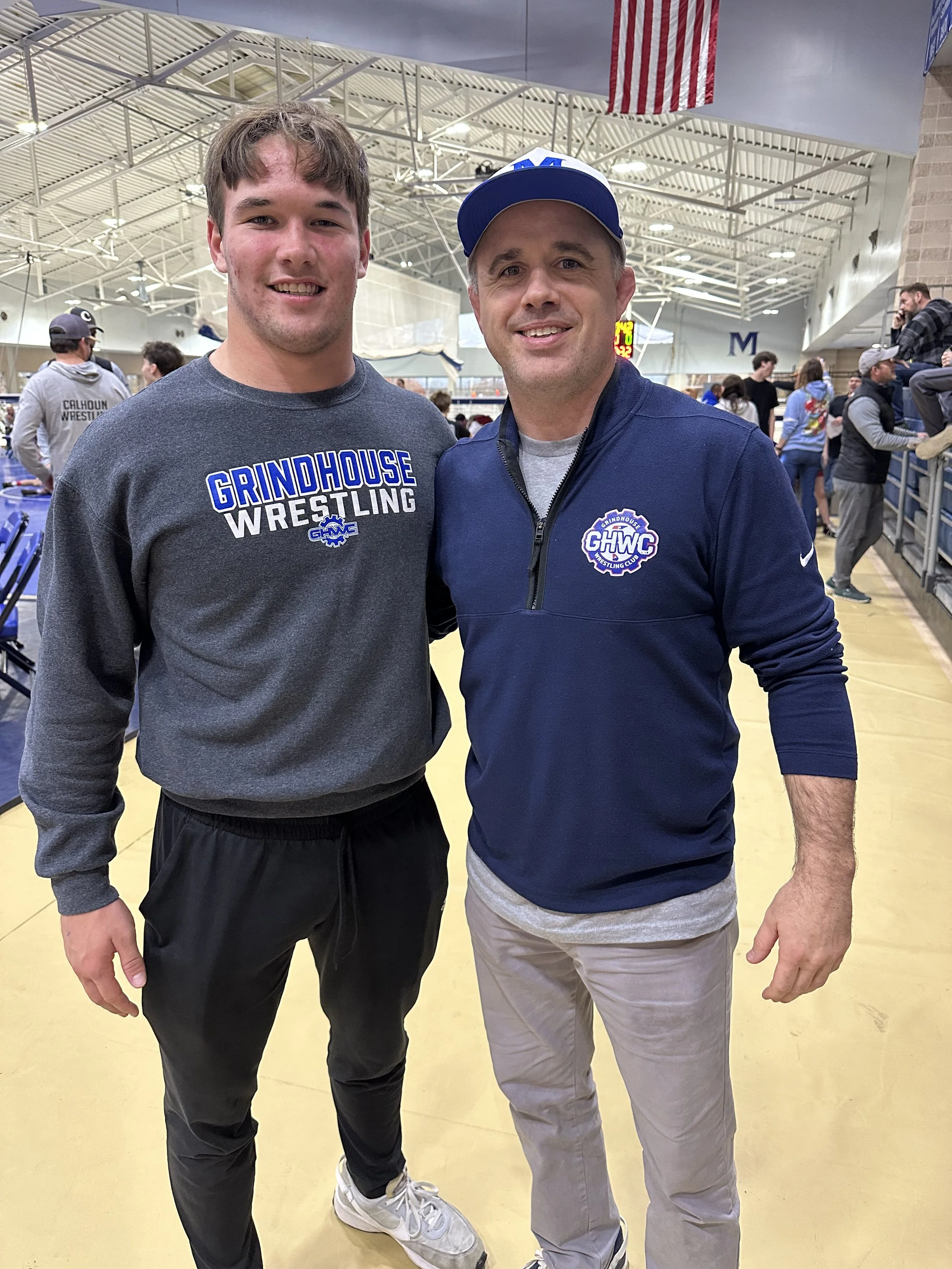 Two young men standing together in a gymnasium, smiling for a photo. The young man on the left is wearing a gray sweatshirt with the words 'GRINDHOUSE WRESTLING' printed on it. The man on the right is wearing a blue jacket with a logo and a cap. There are other people in the background, some wearing wrestling gear, and a large American flag hanging from the ceiling.