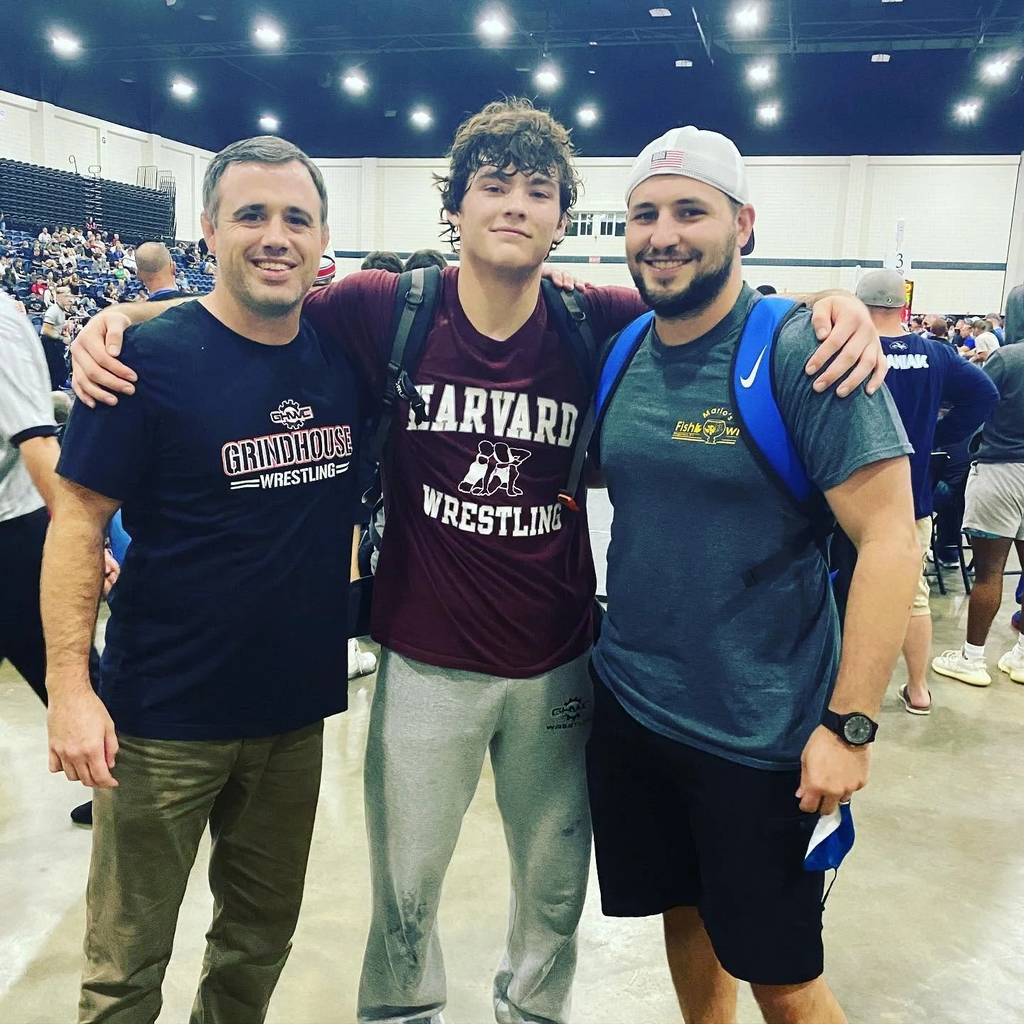 Three men standing together in a gymnasium with a wrestling event happening in the background. They are smiling and have their arms around each other's shoulders. The man on the left wears a black T-shirt that says 'Grindhouse Wrestling', the man in the middle wears a maroon Harvard wrestling shirt and gray sweatpants, and the man on the right wears a gray T-shirt, black shorts, a white baseball cap, and a blue backpack.