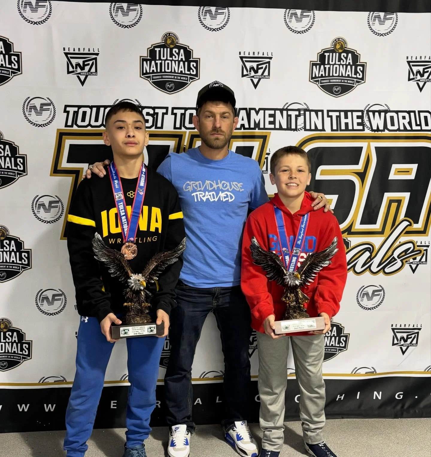 Two boys holding trophies and medals, standing with a man at an indoor wrestling award event. The background features logos and banners for Tulsa Nationals wrestling tournament.