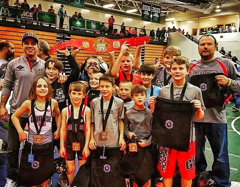 Youth wrestling team with medals on their necks, holding sports gear, and posing with coaches, inside a gymnasium after a tournament.