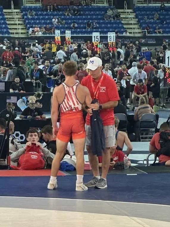 A wrestler in an orange and white singlet stands on a wrestling mat engaging in a conversation with a coach dressed in a red polo shirt, khaki shorts, a white cap, and glasses, at a busy indoor competition with many people and other matches in the background.