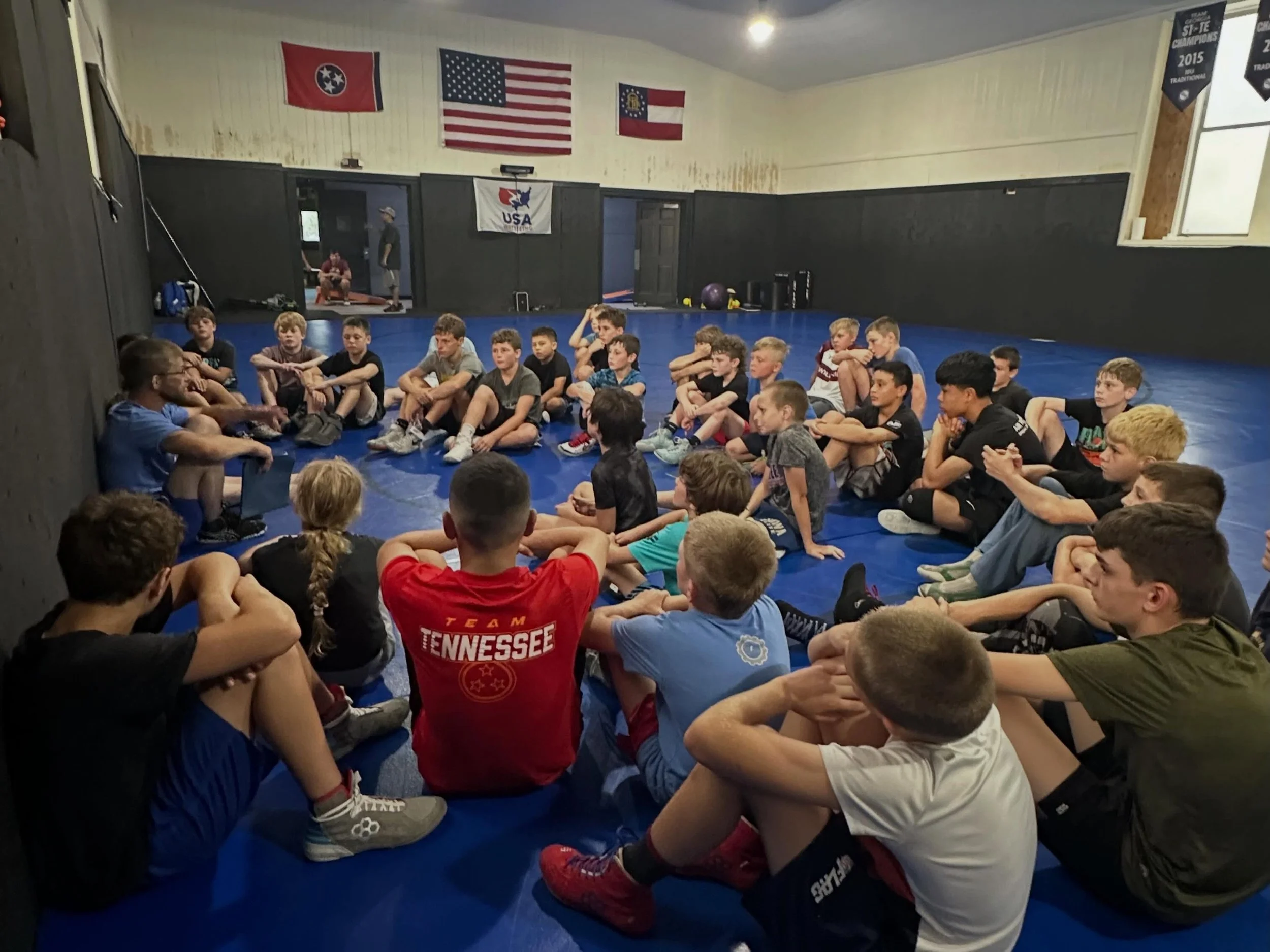 A group of young boys sitting cross-legged on a blue gym mat in a gymnasium, listening to an instructor. The gym walls display flags of Tennessee, the USA, and Georgia, along with banners and gym equipment.