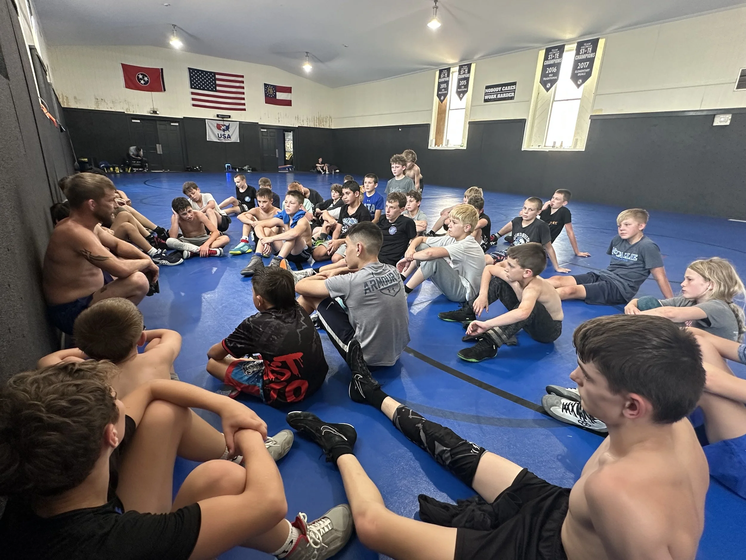 A group of boys in athletic clothing sitting on the blue wrestling mat inside a gym, listening to an adult male coach or trainer.