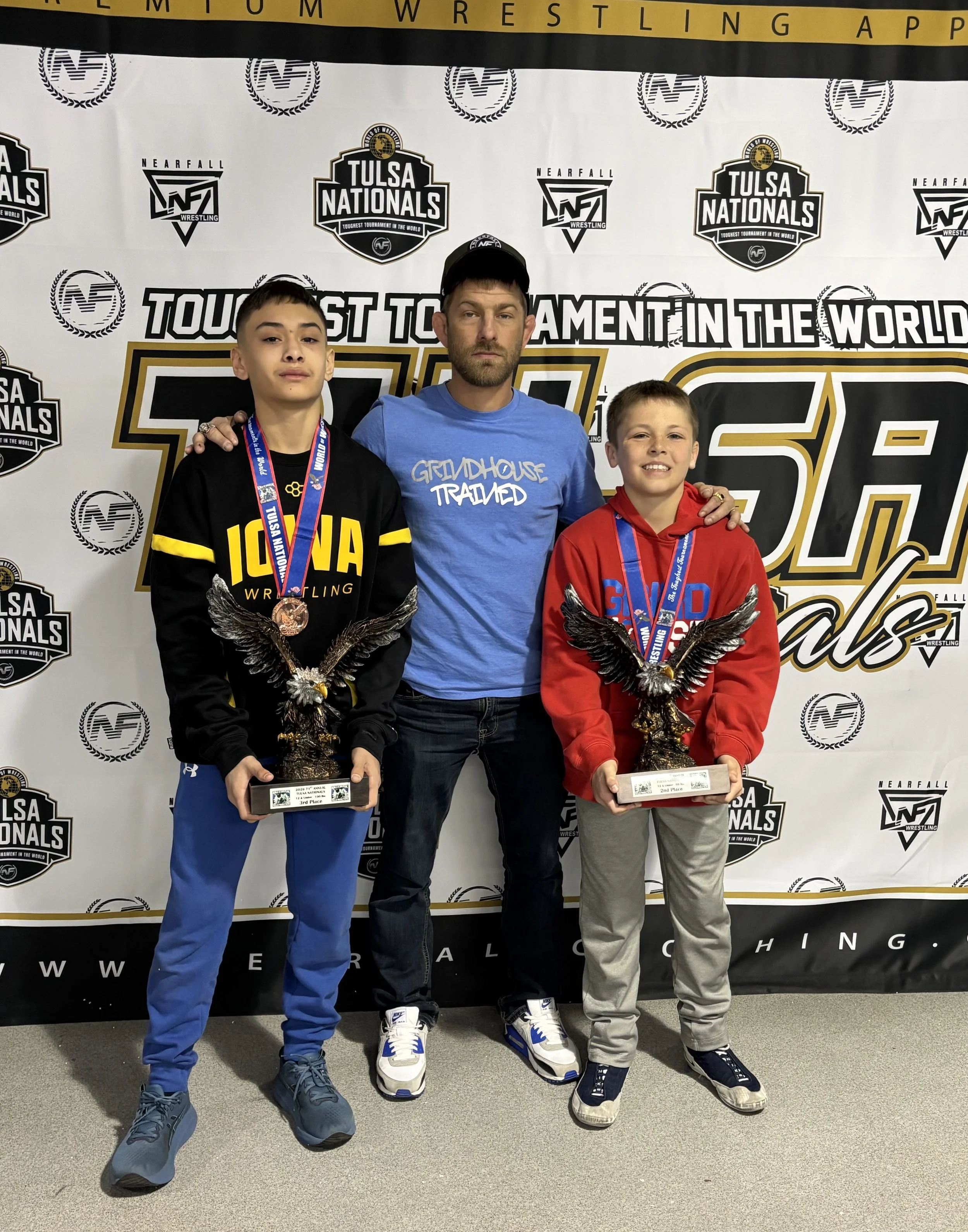 Two young boys wearing medals and holding eagle trophies, standing with an older man in front of a backdrop with wrestling event logos. The boy on the left is wearing a black sweatshirt with yellow lettering, and the boy on the right is in a red hoodie. The man is in a blue T-shirt and a baseball cap.