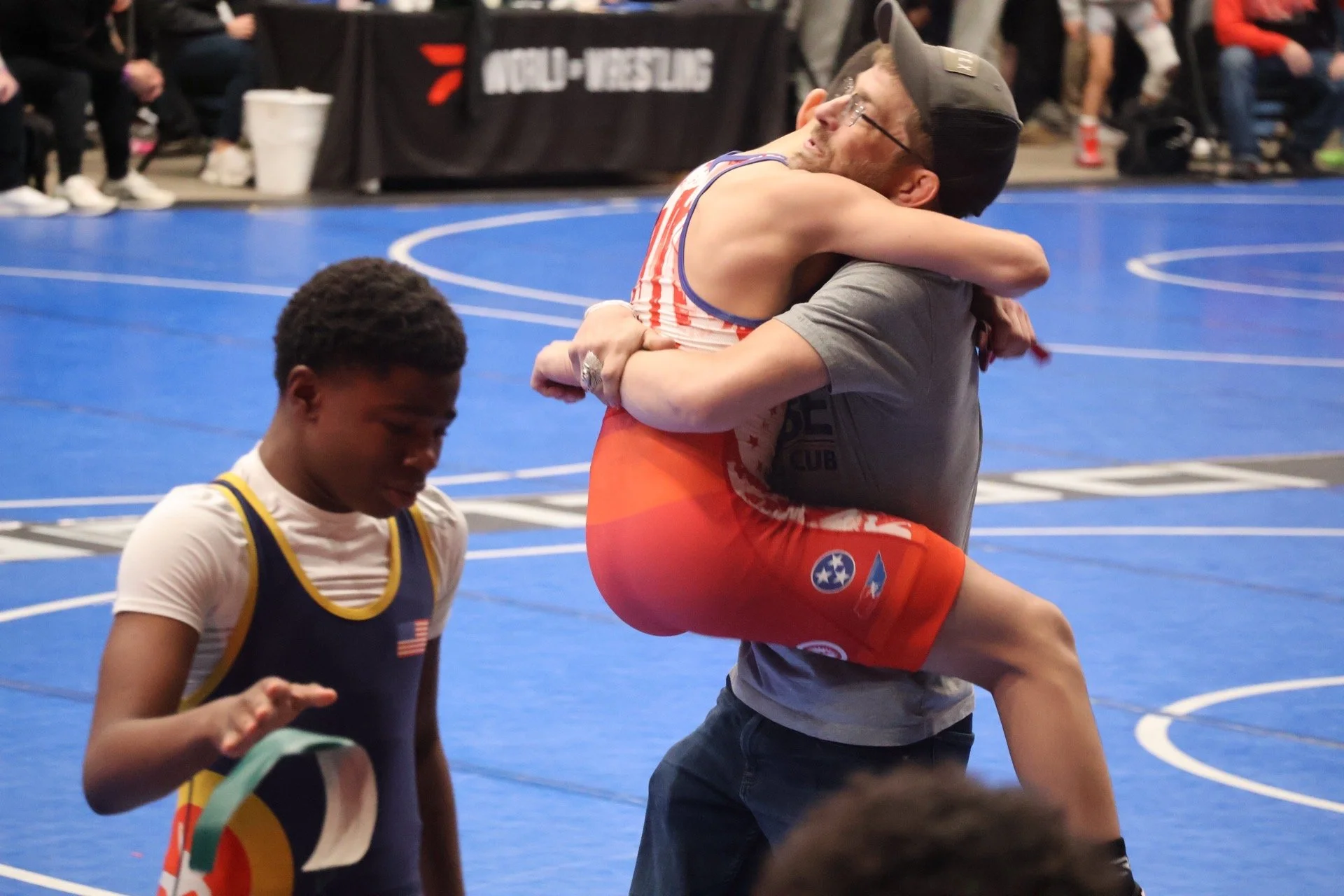 Two wrestlers embrace after a match on a blue wrestling mat, with spectators in the background. One wrestler wears red shorts and a gray t-shirt, the other wears a black singlet with yellow trim.