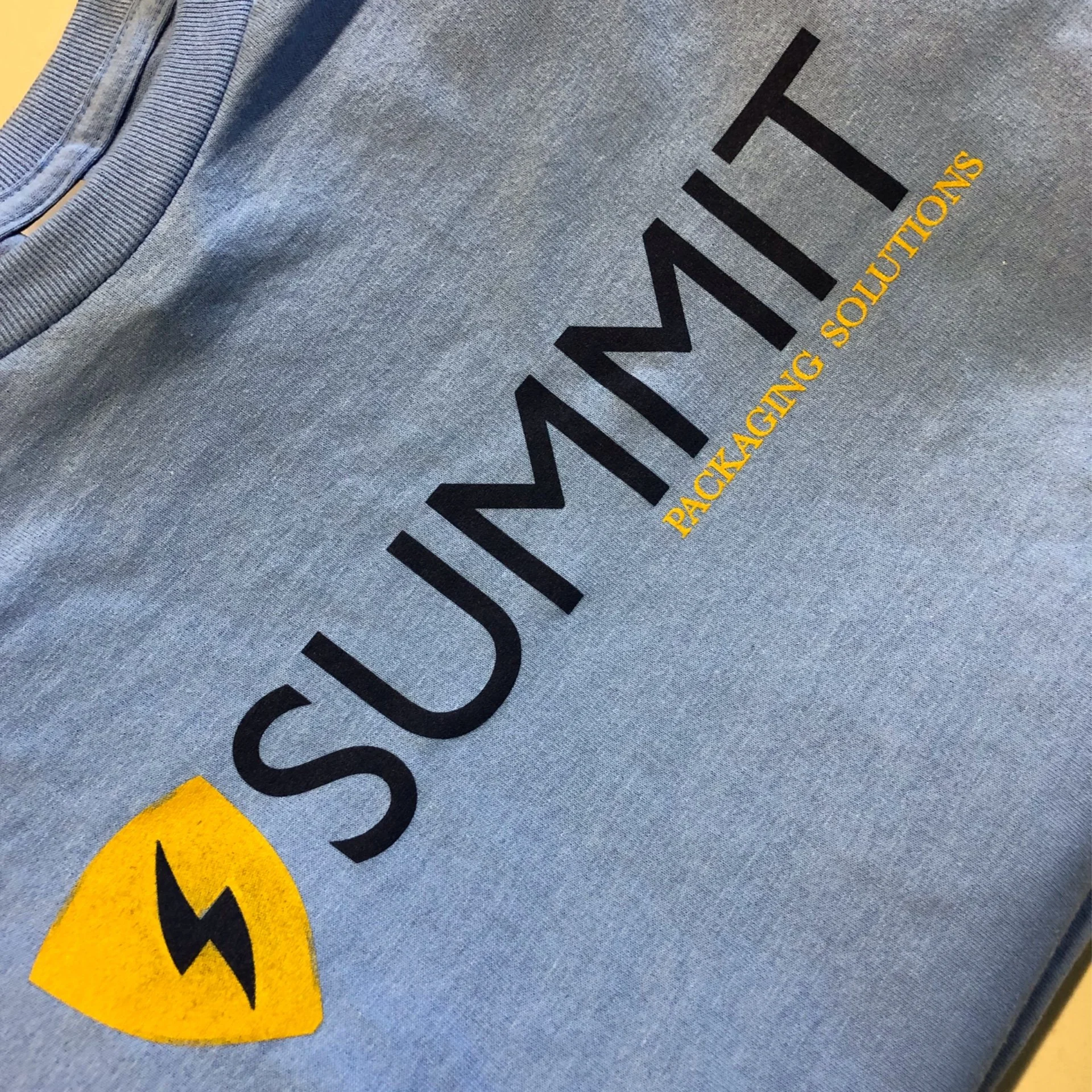 Close-up of a grey hoodie with the word 'Visa' written in black and the phrase 'PACKAGING SOLUTIONS' in yellow underneath. Part of a yellow logo or symbol is visible at the bottom left corner of the image.