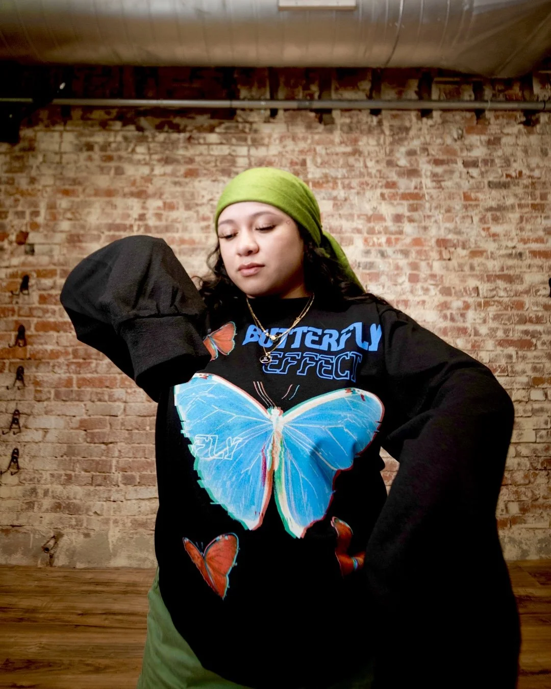 A young woman wearing a green headscarf, black sweatshirt with a large blue butterfly graphic, and olive-green pants, standing in front of an exposed brick wall in a studio.