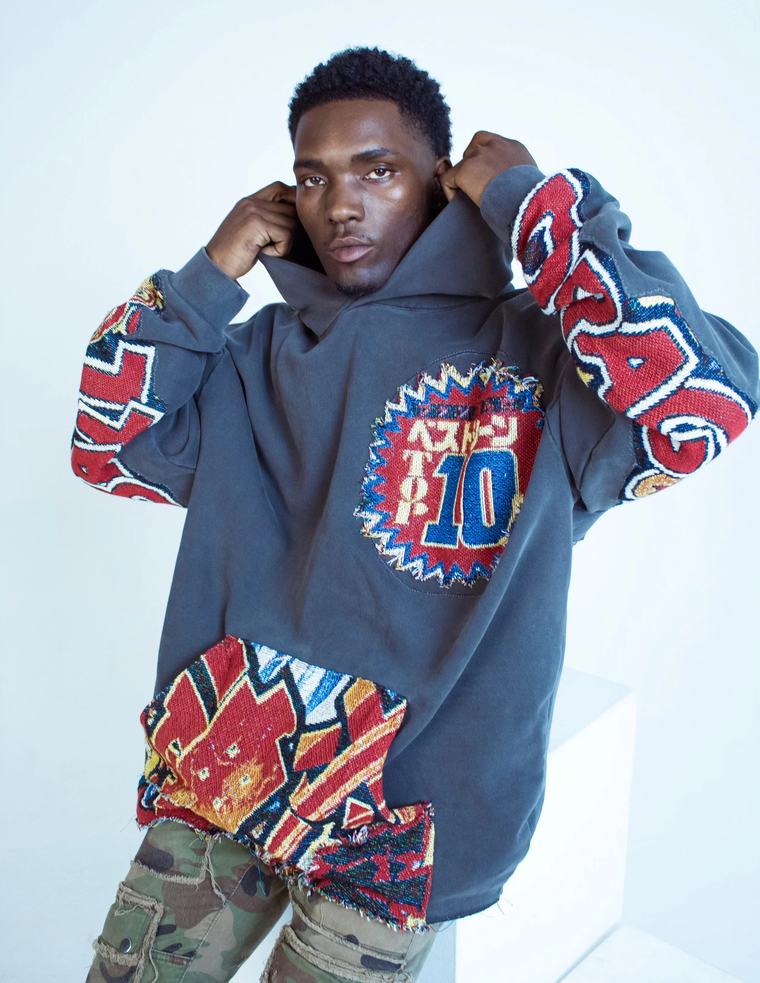 Young man with dark skin and curly hair adjusting the hood of his gray sweatshirt, which has colorful embroidered patches on the sleeves and chest, paired with military camouflage pants.