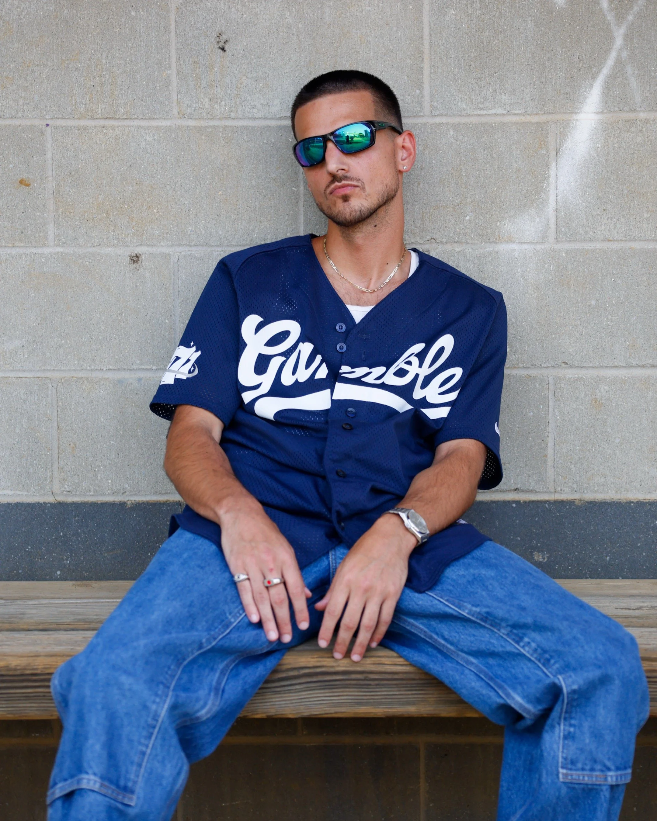 Young man with short dark hair, wearing mirrored sunglasses, a blue sports jersey with white lettering, several rings, a bracelet, and a necklace, sitting on a wooden bench against a cinder block wall.