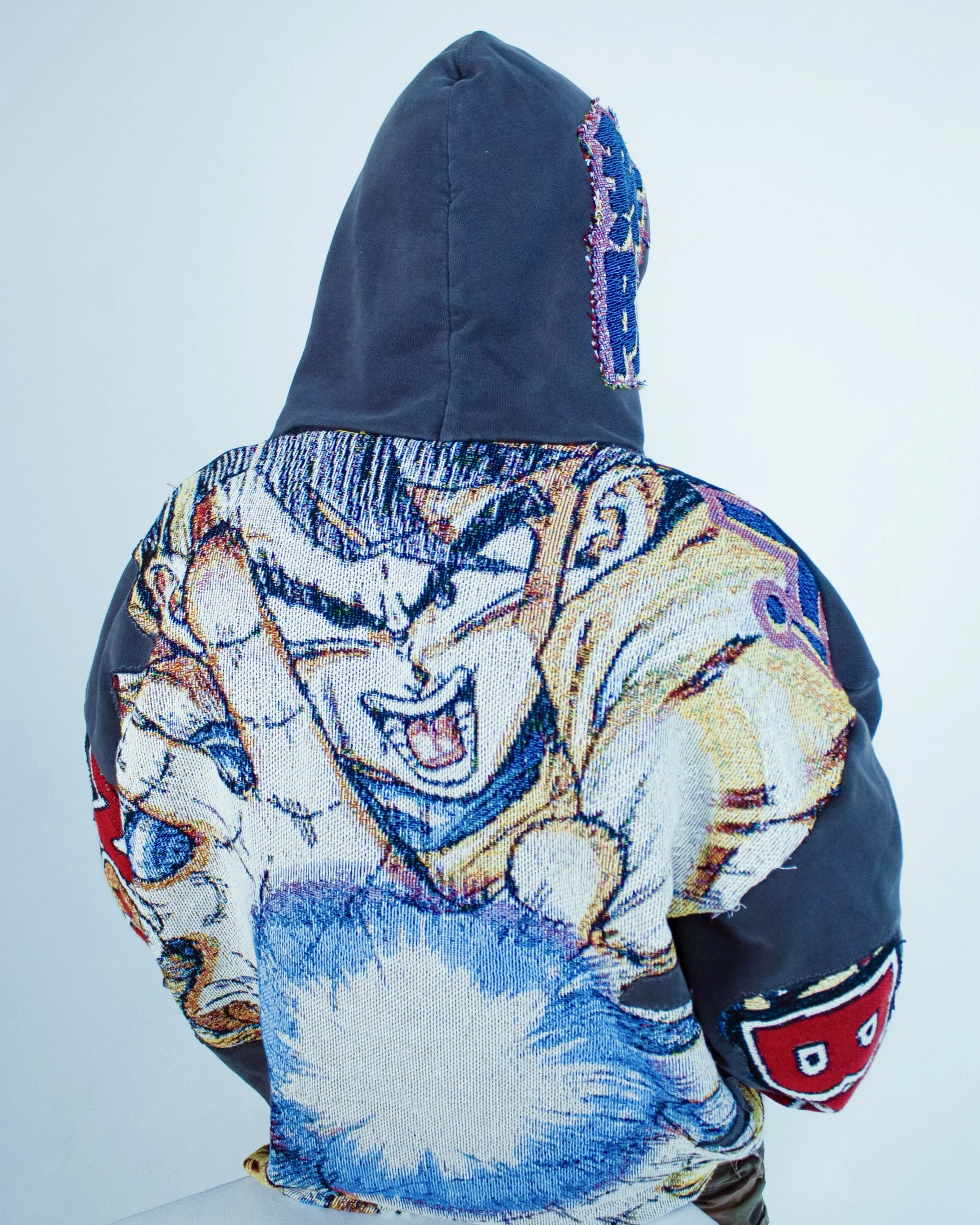 Back view of a person wearing a black hoodie with a colorful embroidered patch on the back, mixed with artwork of a tiger and abstract patterns; short dark hair, light gray background.