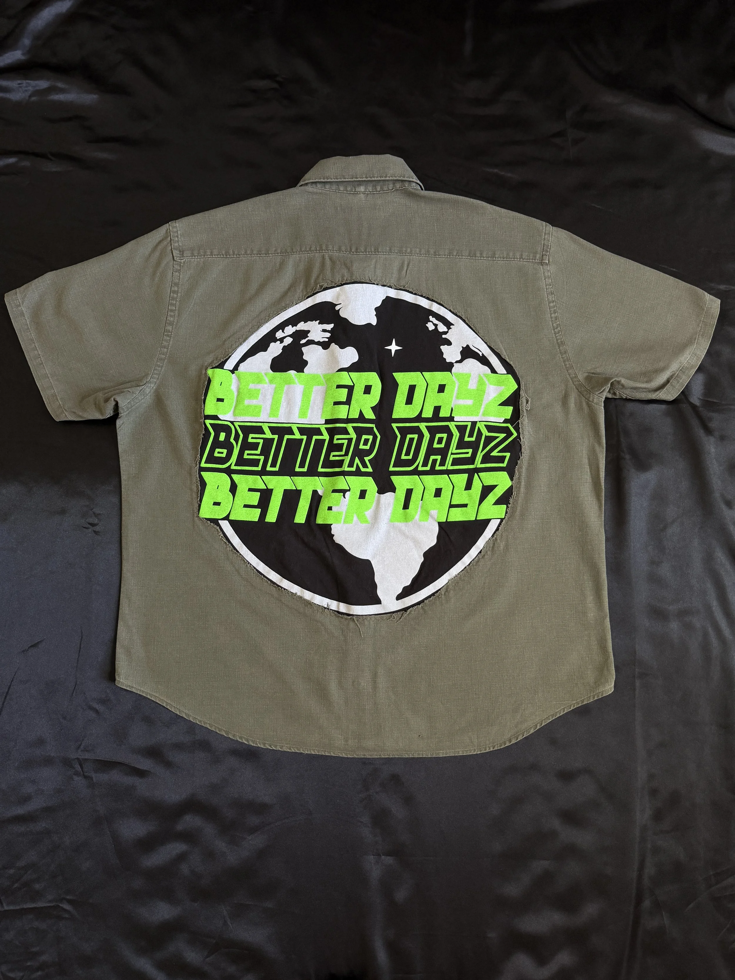 A tan short-sleeve button-up shirt with a large circular patch on the back featuring a black and white world map and the repeated phrase 'BETTER DAYZ' in bright green text.