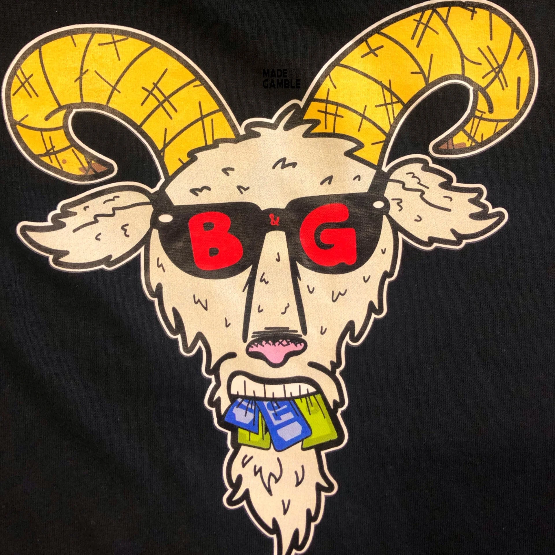 Stylized illustration of a ram's head with large yellow horns marked with black lines, wearing black sunglasses with red lenses, red lips, a fluffy beige face, and blue and yellow shorts with white lettering.
