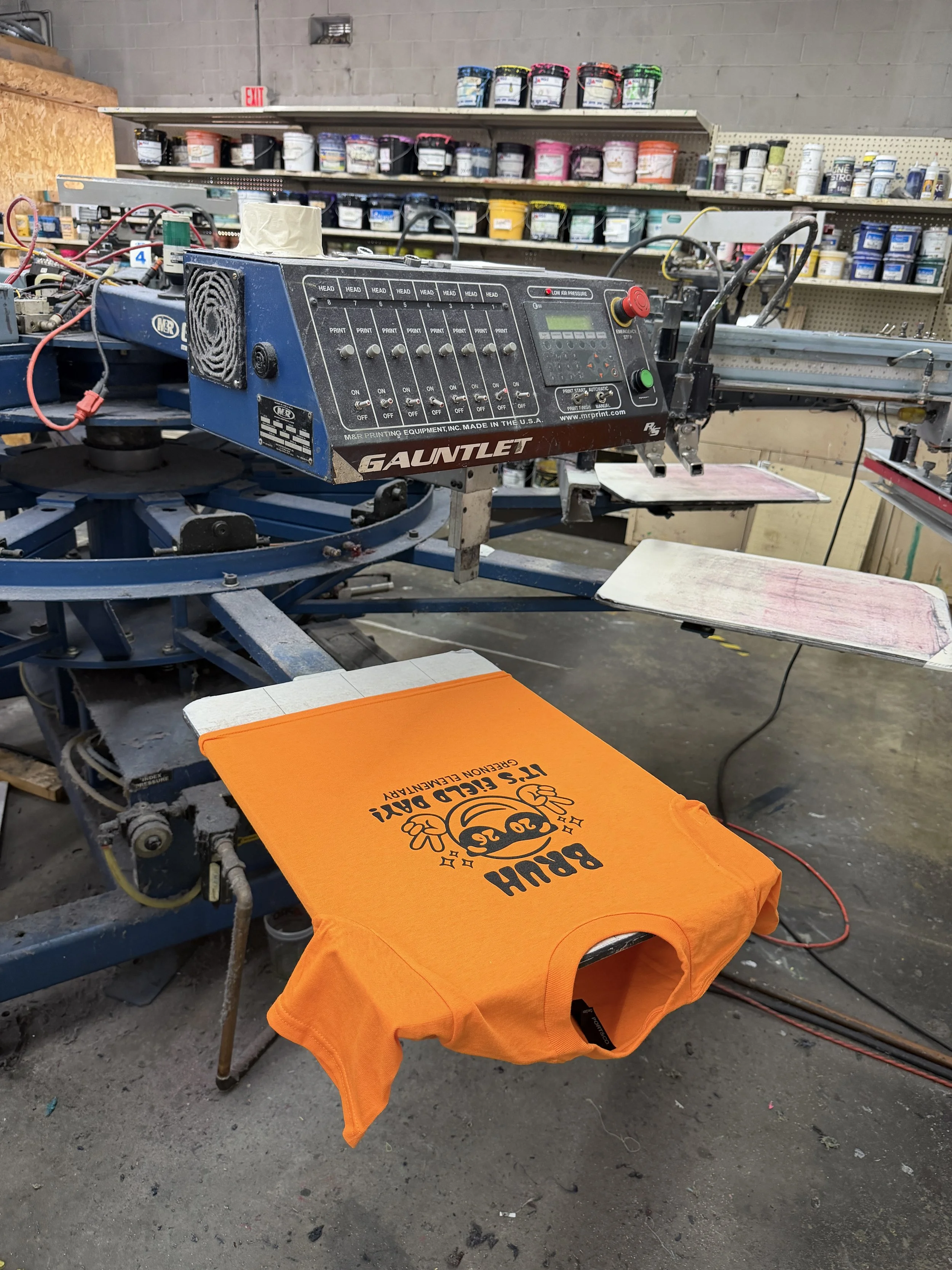 A printing press machine on a work table in a workshop, with an orange T-shirt with the words 'BLOOM' and a graphic of a flower on it laid out on the table.