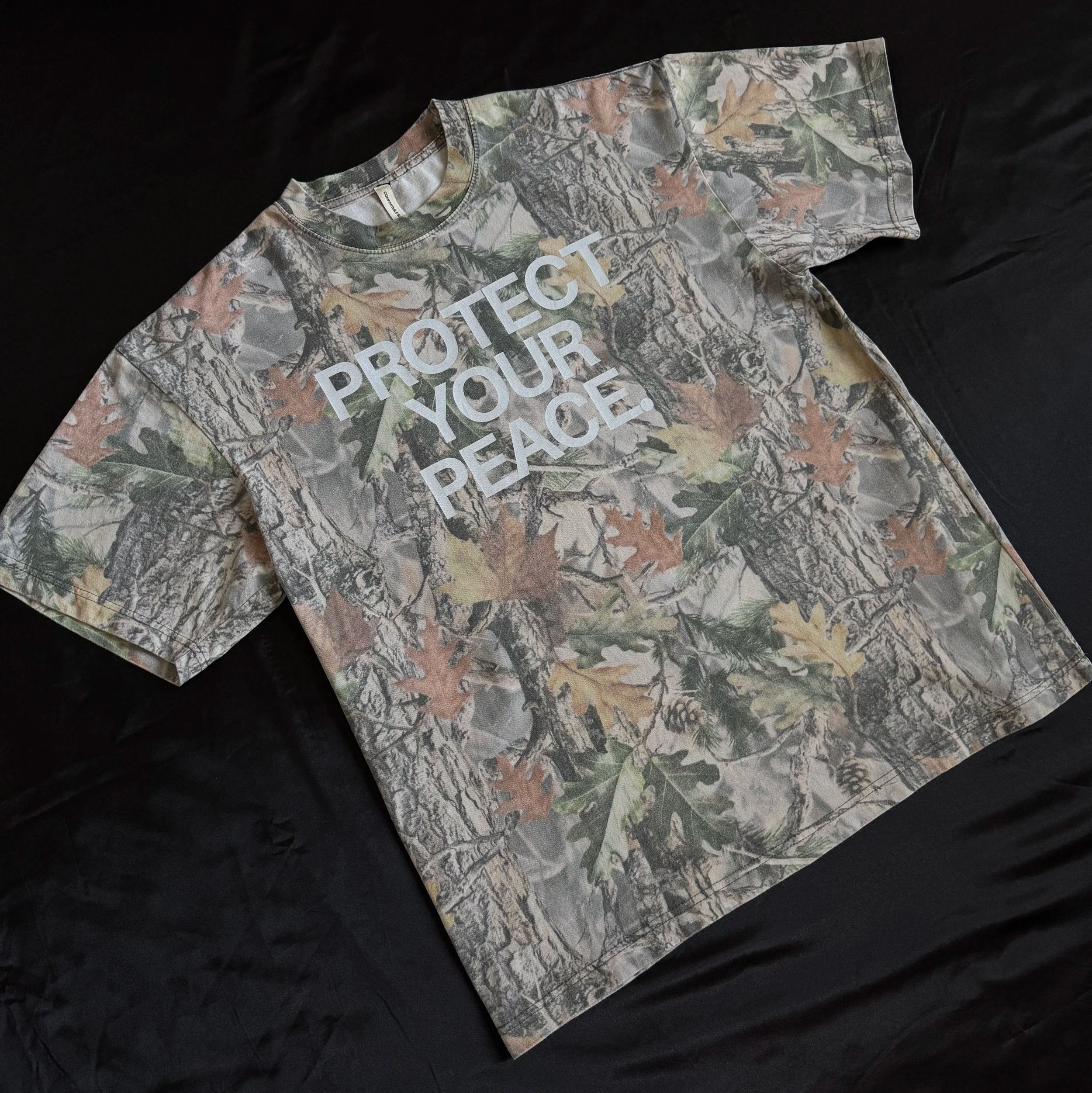 Camouflage T-shirt with white text reading 'PROTECT YOUR PEACE' lying on a black background.