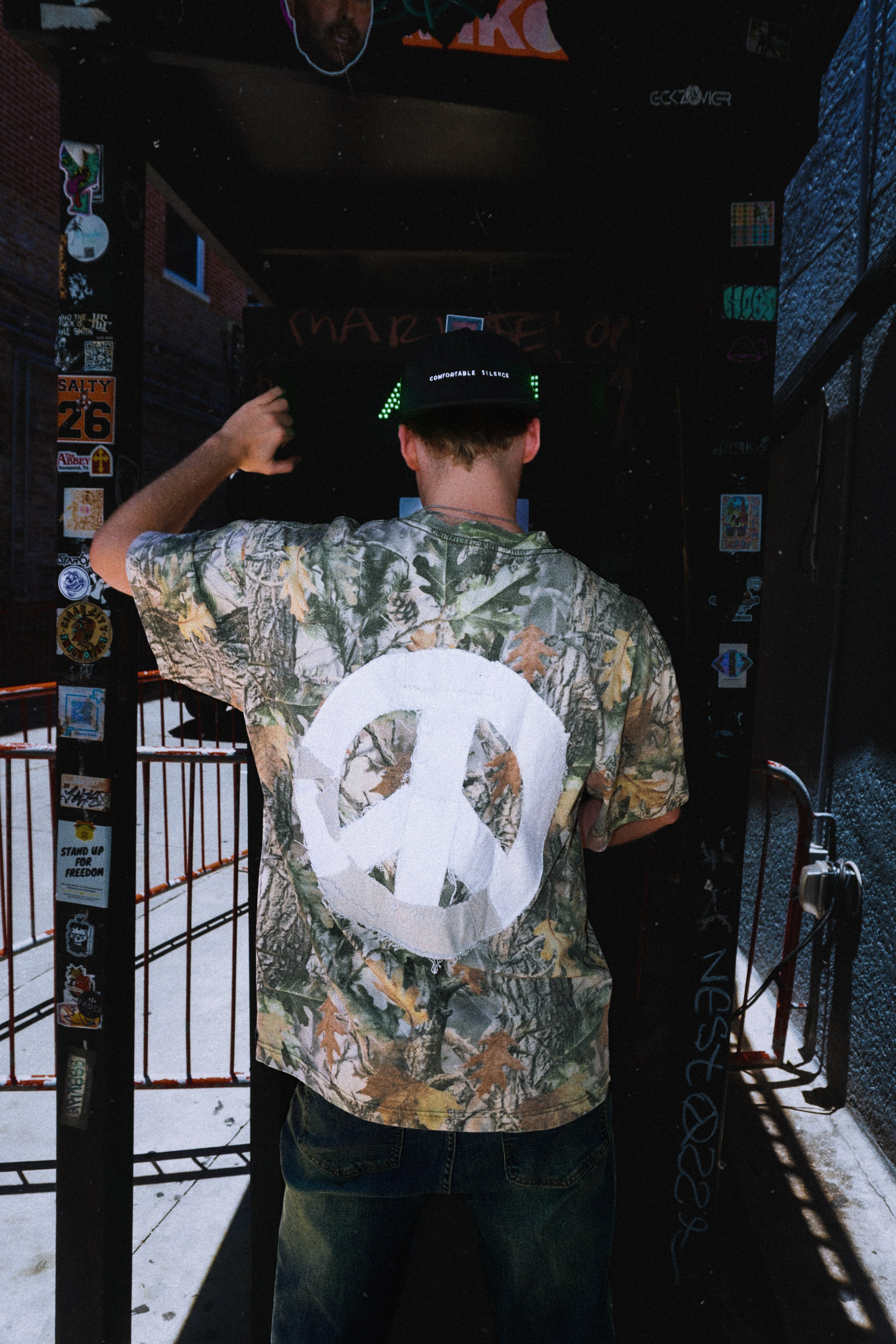 Person wearing a camouflage shirt with a large white peace symbol on the back, adjusting a black hat with the words 'Comfortable Silence' on it, standing in front of a dark door with stickers and graffiti.