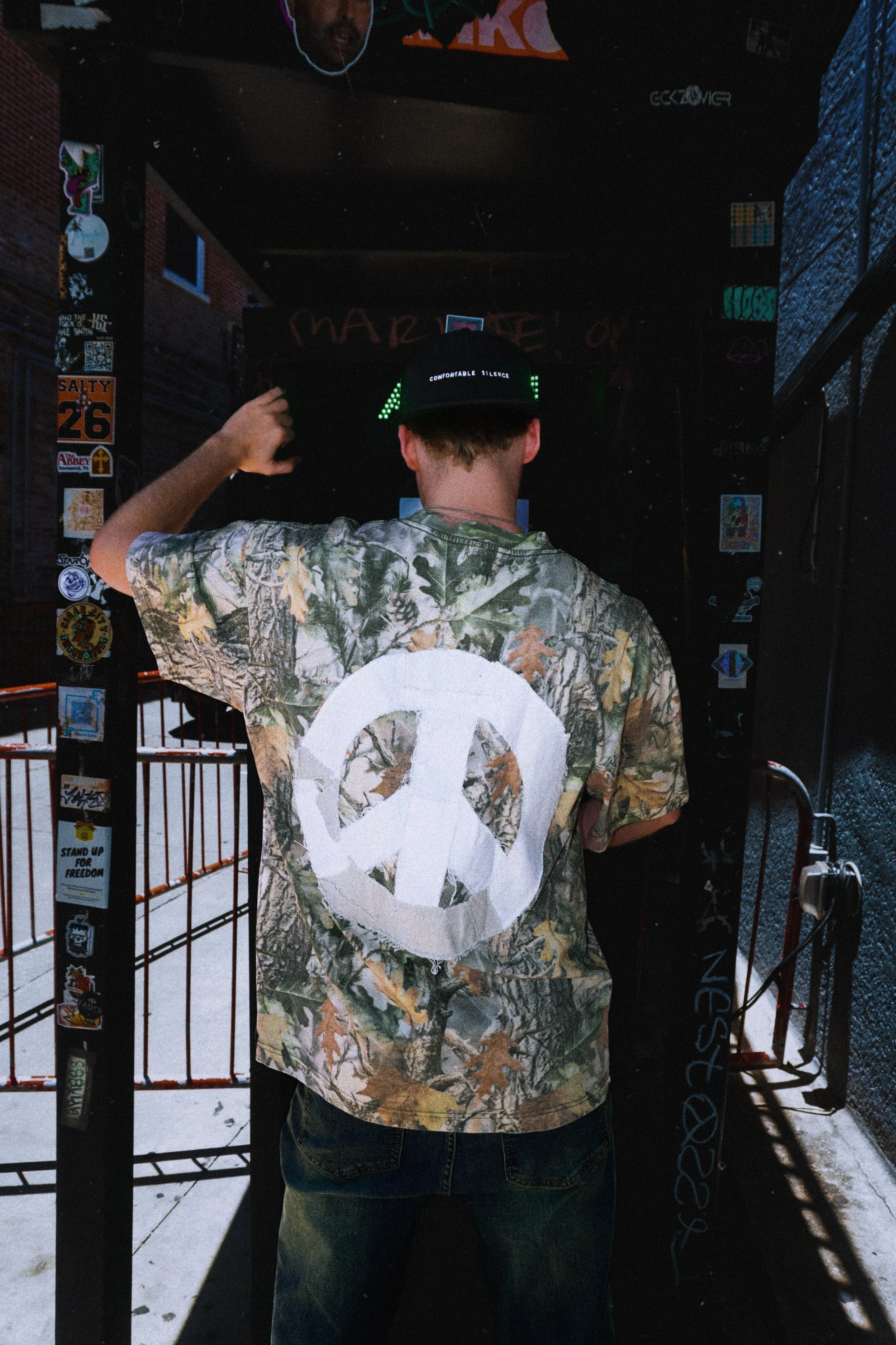 Person facing away wearing a camouflage shirt with a large white peace symbol on the back, black cap with white text, standing at a dark entrance with various stickers and graffiti, outside in snow.