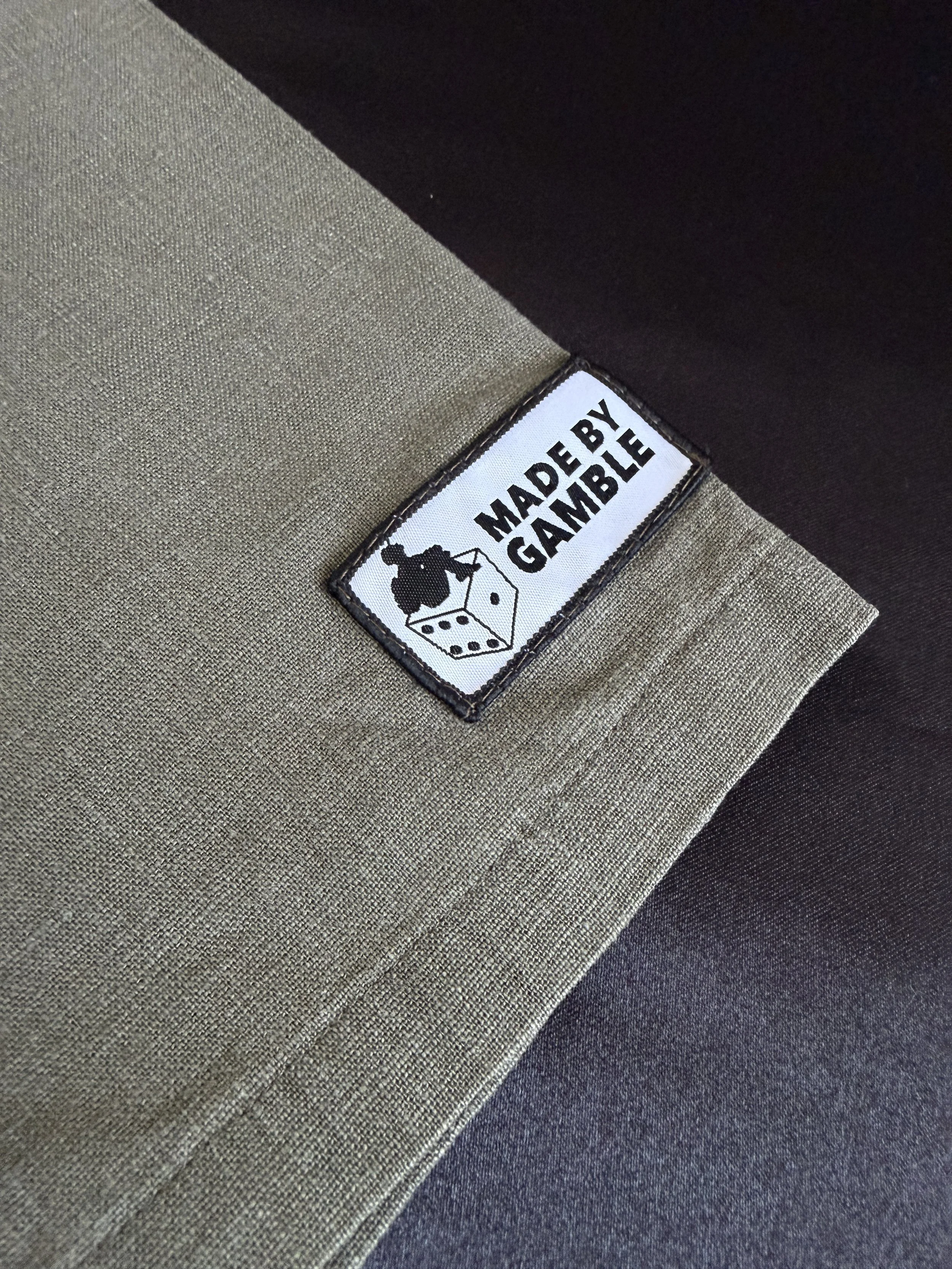 Close-up of a fabric label on a piece of clothing that reads 'Made by Gamble' with a silhouette of a person sitting on a box. The clothing appears to be made of beige and dark gray fabric.