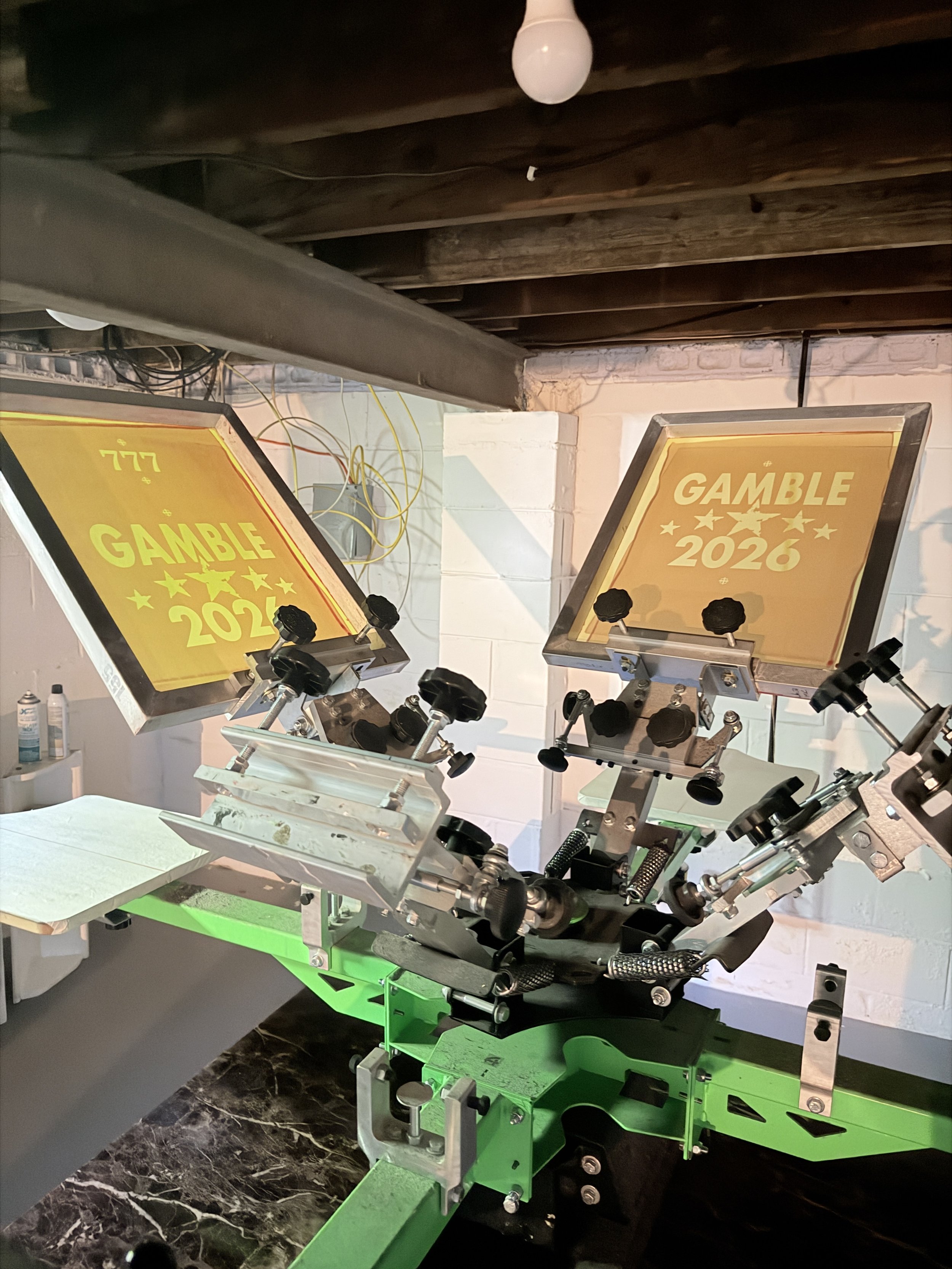 A mechanical device with various knobs and parts, placed on a green stand, with two illuminated screens displaying the text "GAMBLE 2026" and a yellow background with stars, in a room with a wooden ceiling and exposed wiring.