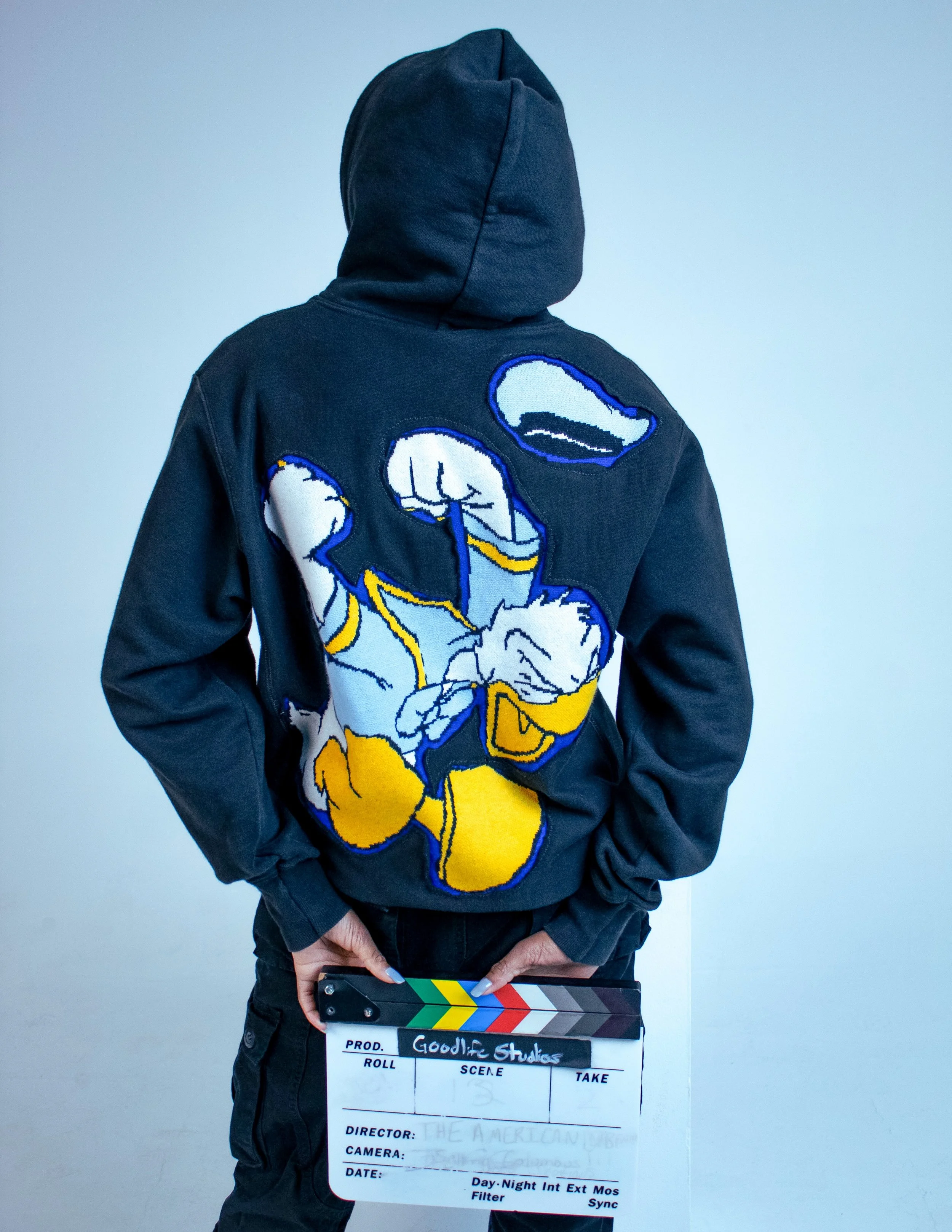 Person wearing a black hoodie with a colorful cartoon of Donald Duck embroidered on the back, holding a film clapperboard labeled 'GoodLife Studios' in front of a plain gray background.