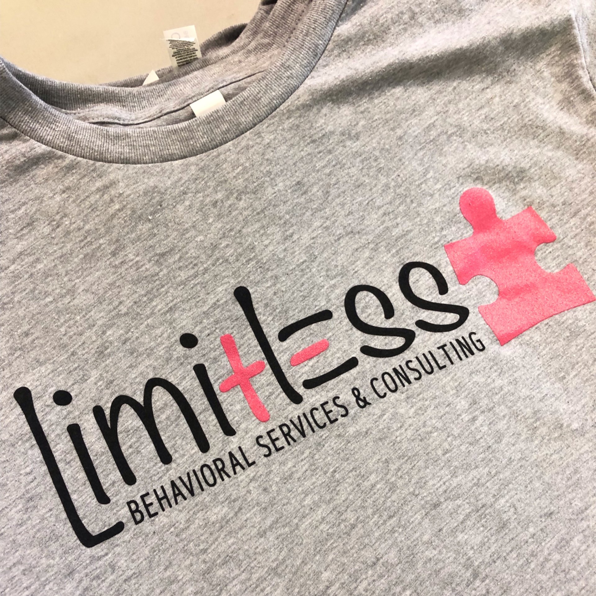 Close-up of a gray T-shirt with a logo and text on the front, including a pink puzzle piece graphic and the words 'Behavioral Services & Consulting'.