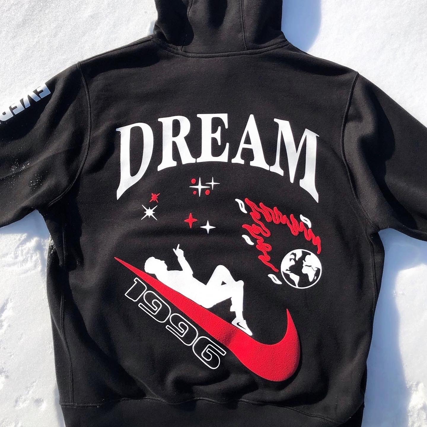 Black hoodie with white, red, and black graphic text and images including the word 'DREAM', a planet Earth, a silhouette of a person, and NASA logo elements on the snow.