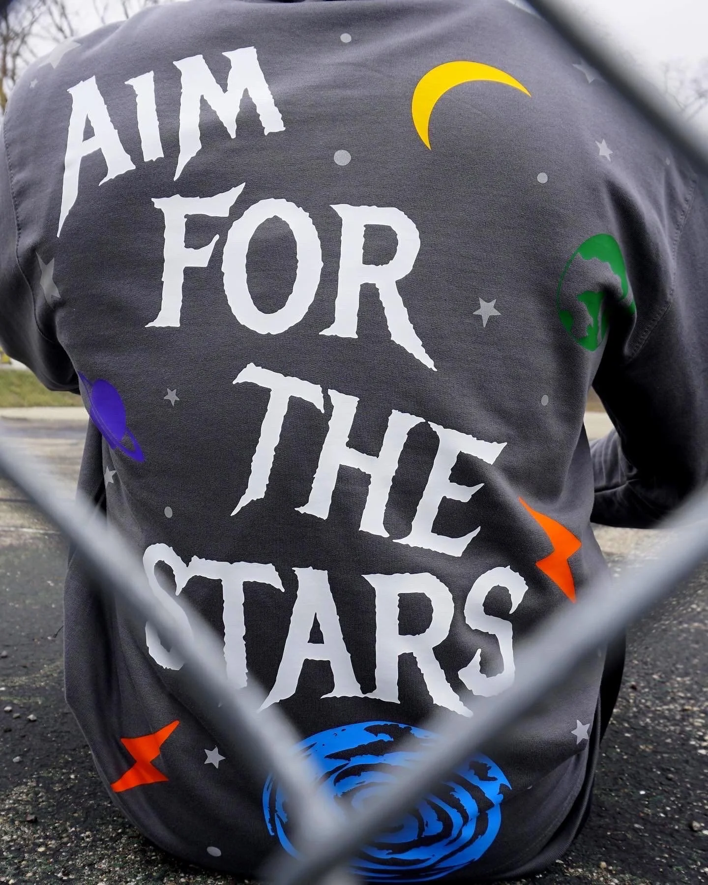 A person wearing a dark grey uniform with the words 'Aim for the Stars' in large white letters on the back, along with colorful star and moon designs, and the image is taken through a fence.