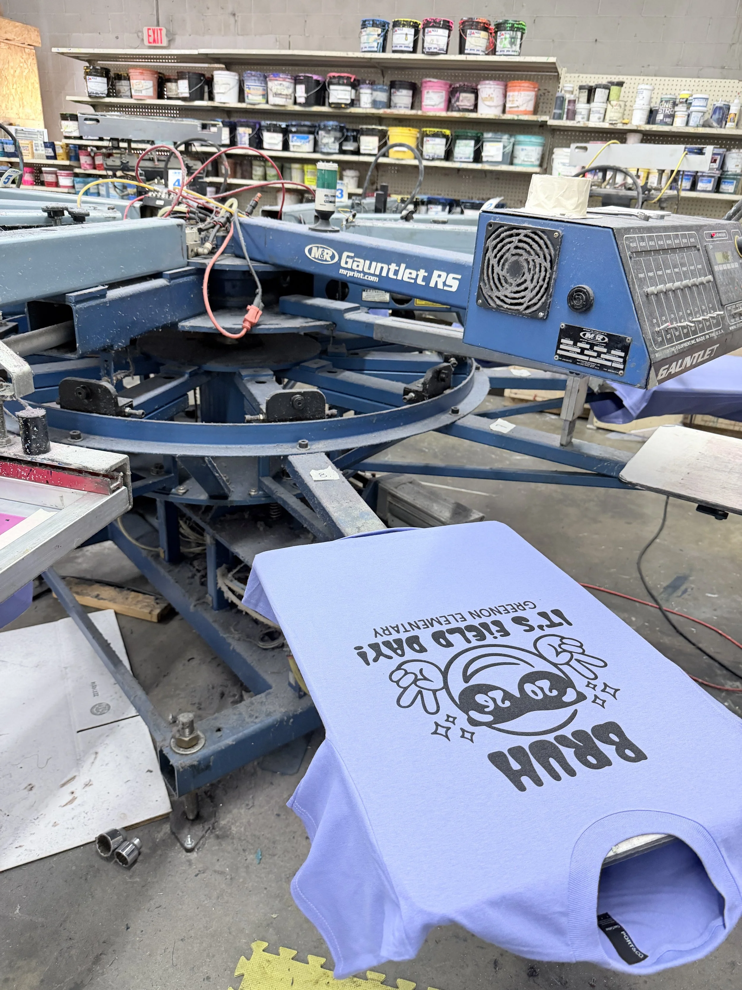 A large screen printing press in a workshop, with a light blue t-shirt that reads 'BIRTHDAY BOY' and features a cartoon face, laid flat beneath the press for printing.