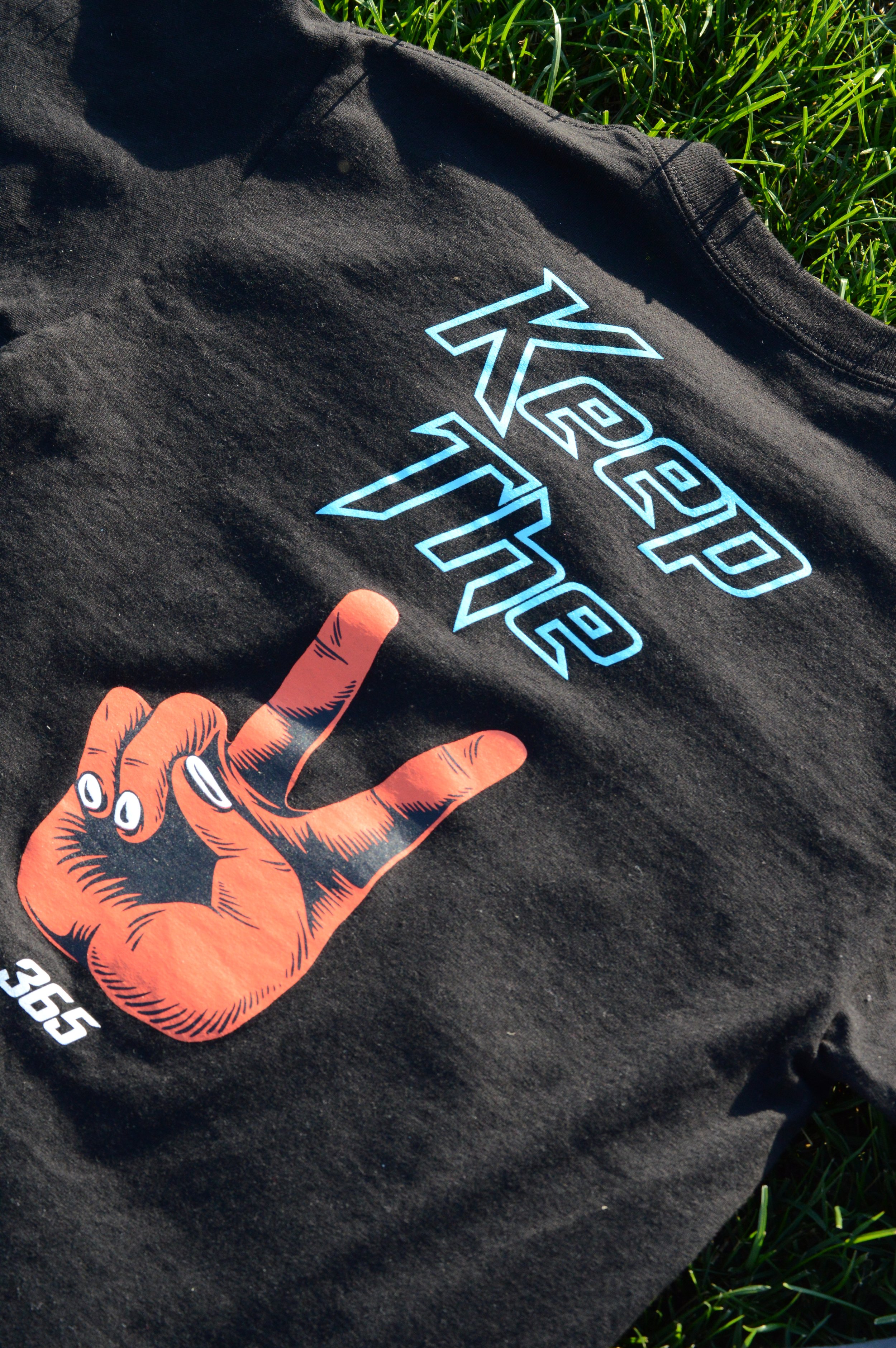 A black T-shirt with a graphic of an orange hand and blue outlined text that says "TIGER" with the word "ROAR" inside the letter "A." The shirt is on green grass.