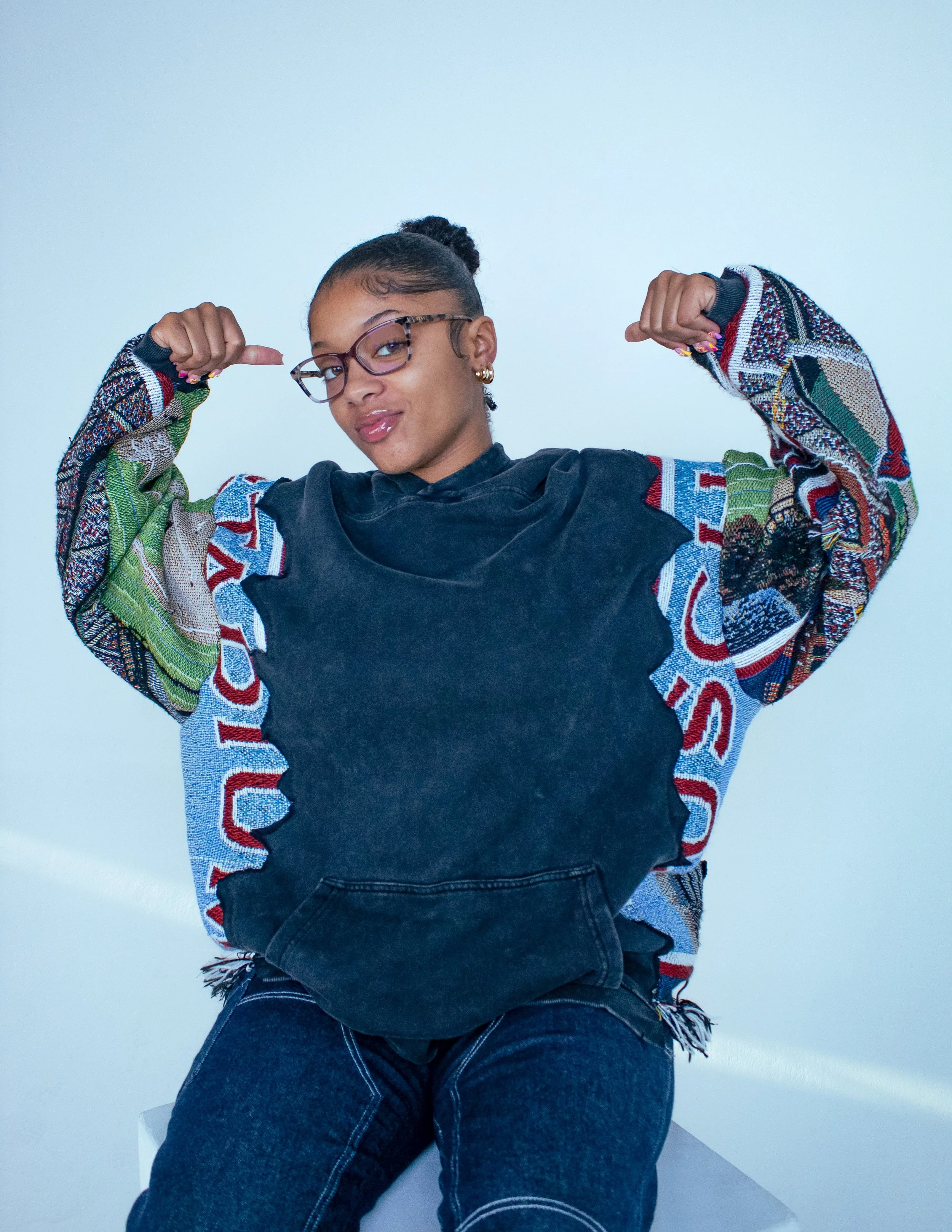 A young woman with dark hair, glasses, and hoop earrings, flexing her arms in a confident pose against a light blue background. She is wearing a black sweatshirt with colorful patterned sleeves and black jeans.