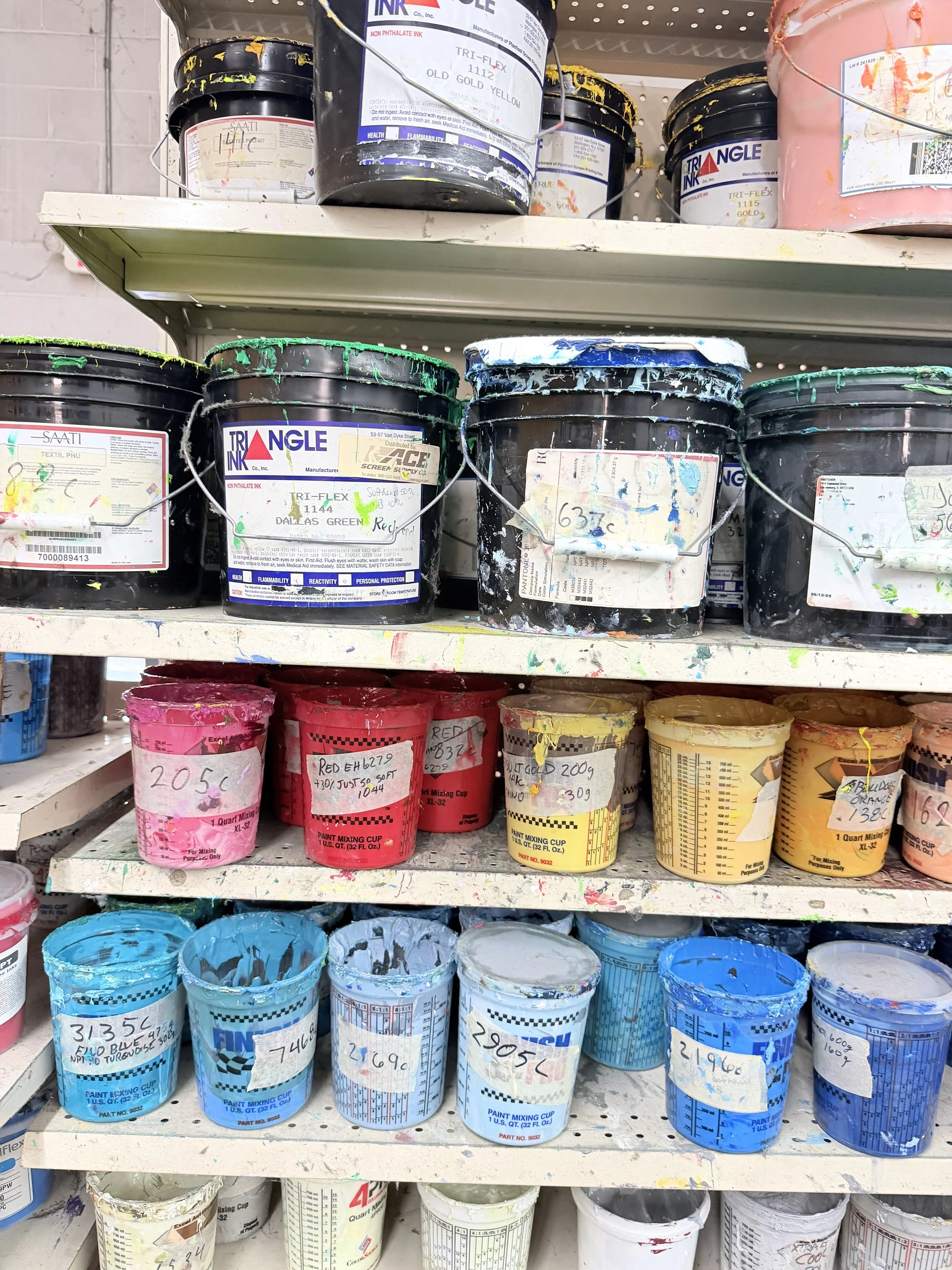 Paint cans on store shelves, including black, pink, red, yellow, and blue buckets, many with visible labels and paint splatters.