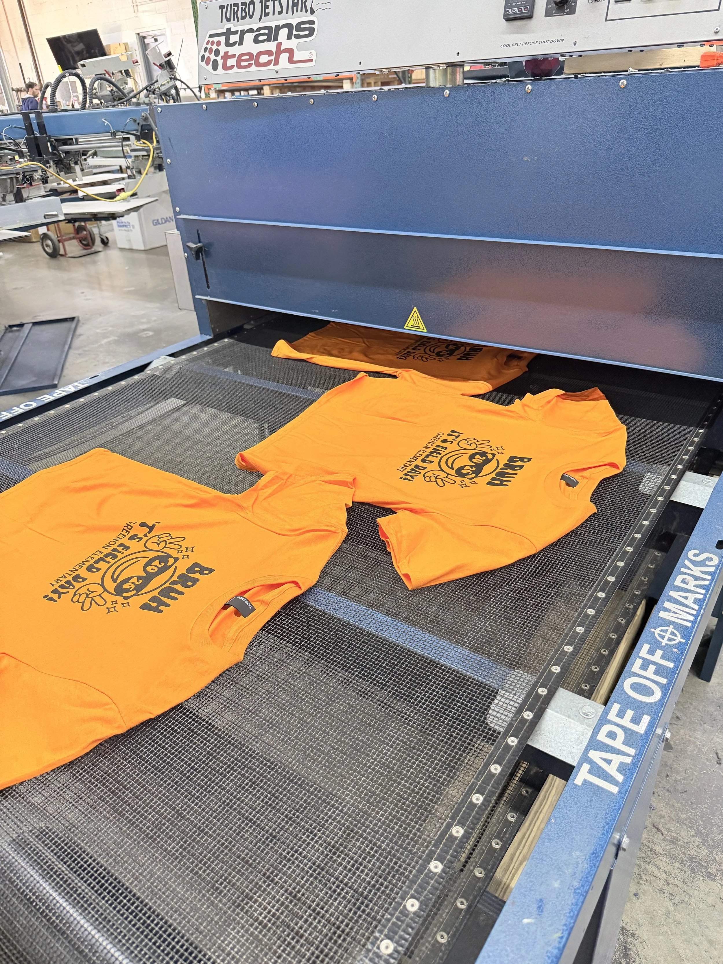 Bright orange T-shirts with black printed graphics are laid flat on a conveyor belt in a manufacturing or printing shop. The T-shirts display the phrase 'Brih Eki' and a cartoonish chicken graphic. In the background, parts of industrial equipment and