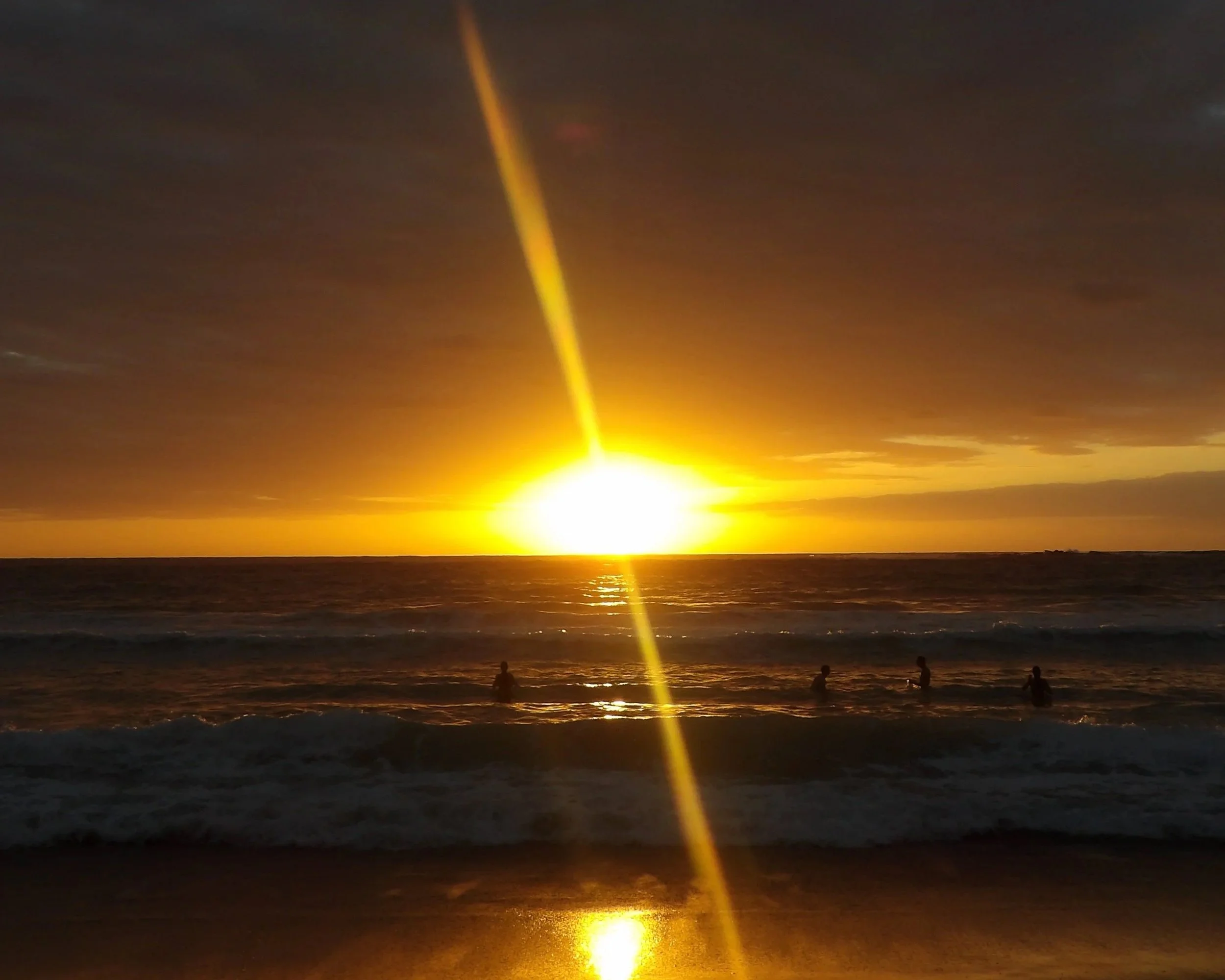 Sunrise over the ocean with a bright yellow beam of light extending from the horizon into the sky, and silhouettes of people in the water.