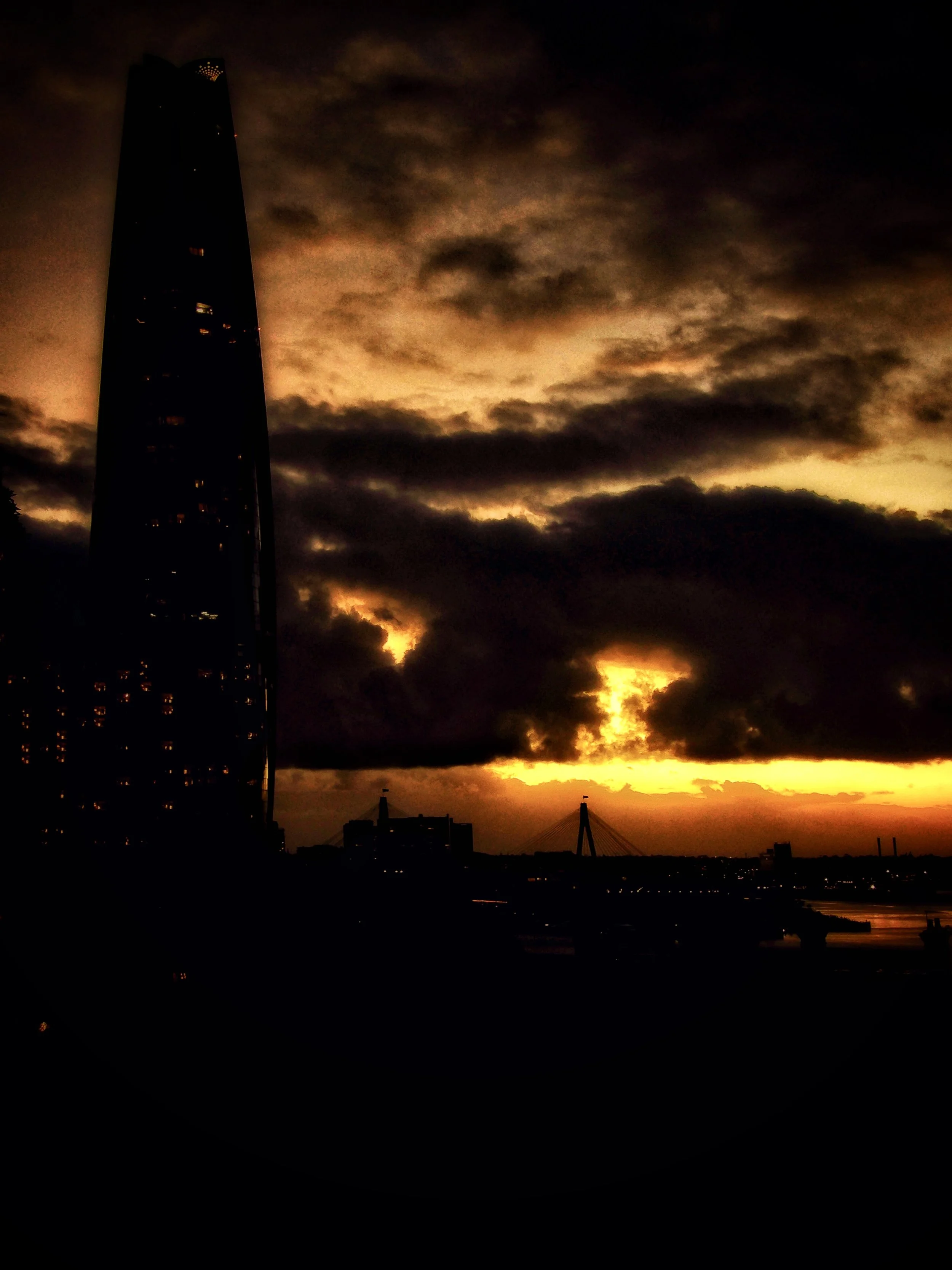 A city skyline at sunset with dark clouds and a tall skyscraper on the left, and a bridge in the distance on the right.
