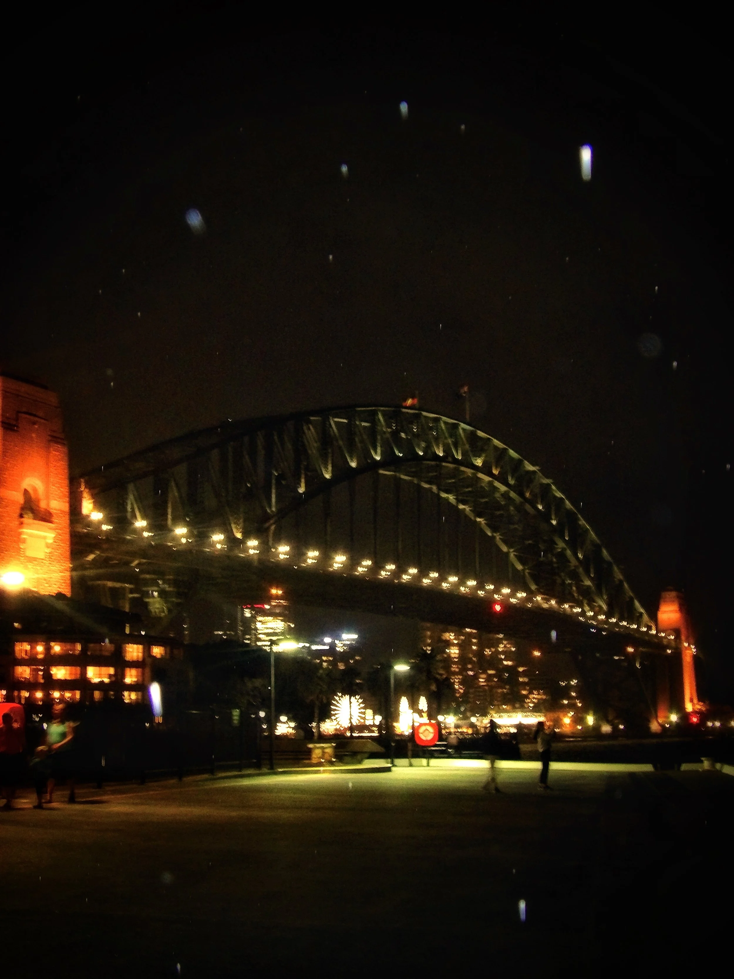 Night view of a bridge with illuminated arches, buildings on the left with warm lighting, a Ferris wheel in the distance, and people walking in the foreground.
