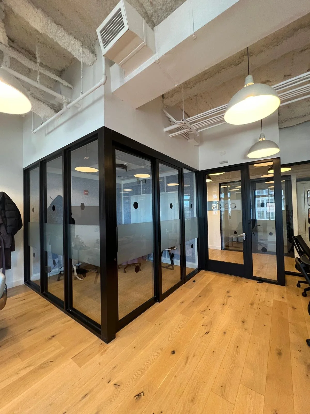 Modern office space with glass-walled meeting rooms, wooden flooring, and exposed ceiling with white pipes and vents, pendant lights hanging from ceiling.