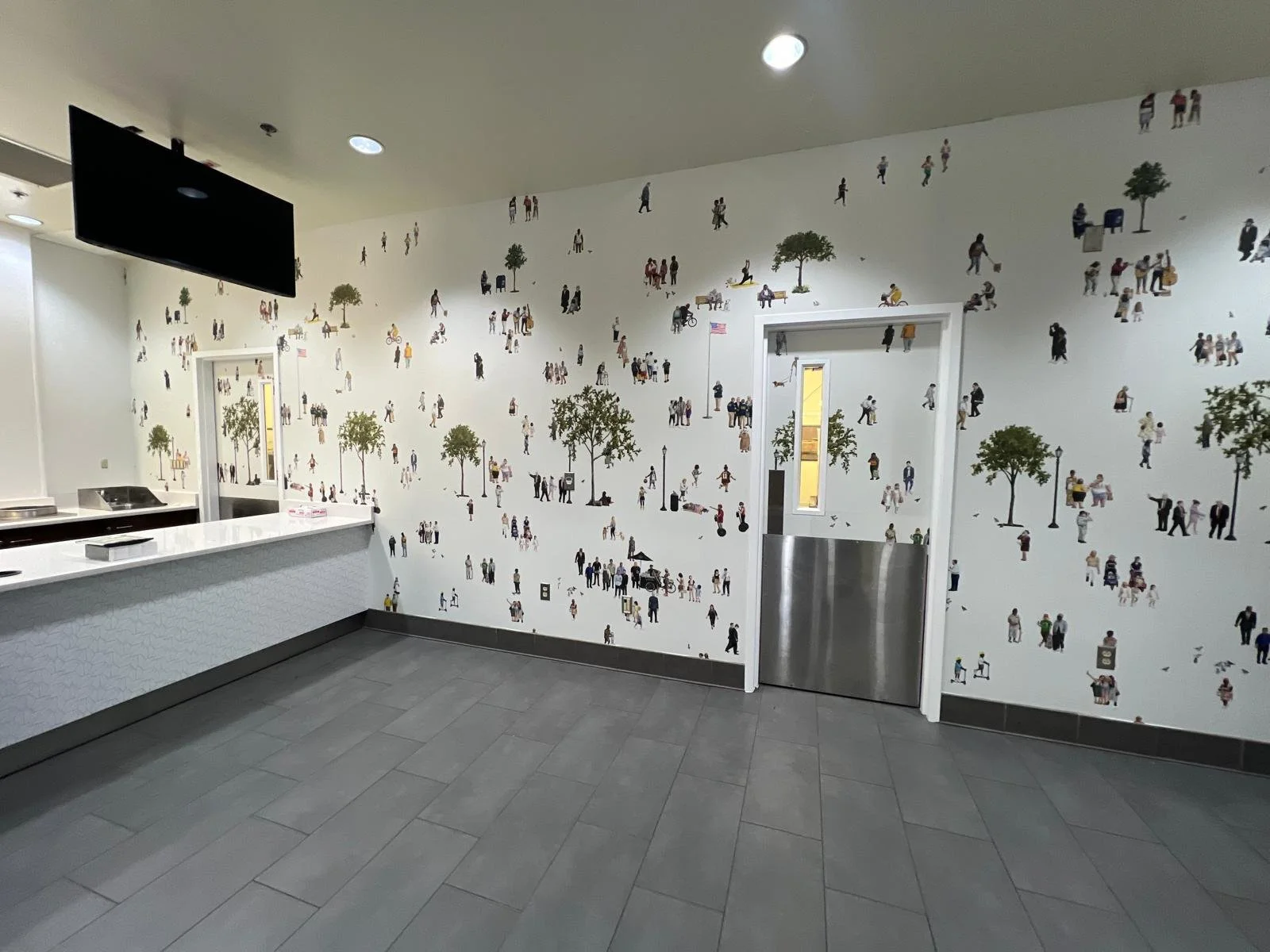 Interior of a building with a white wall decorated with numerous small illustrations of people, trees, and outdoor scenes, resembling a mural or wallpaper. There is a counter on the left side and an open door leading to another room.