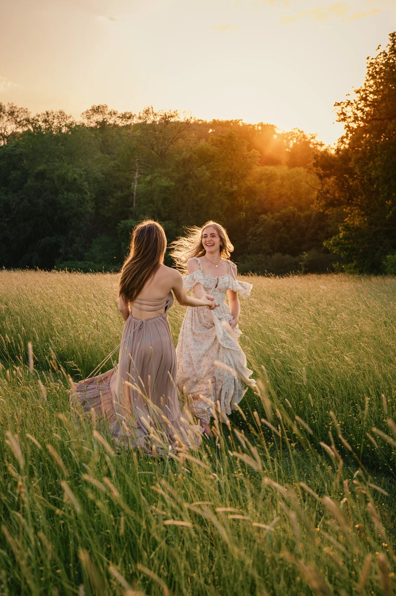Two women in long skirts holding hands and dancing in a grassy field at sunset, with trees in the background.